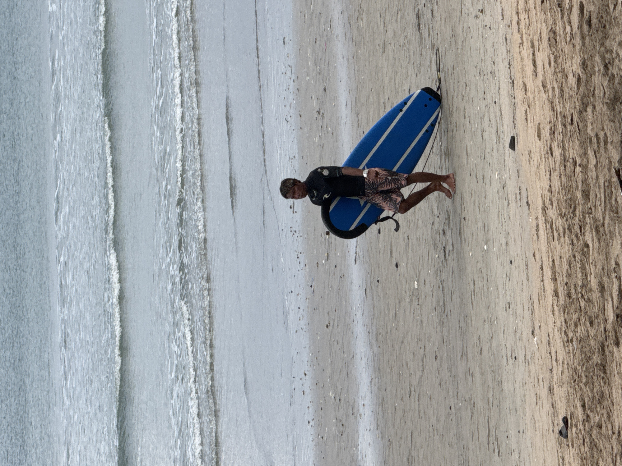 A man is walking on a sandy beach carrying a blue surfboard with white stripes. He is wearing a black wetsuit and patterned shorts. The ocean is in the background with gentle waves. The beach is scattered with small shells and rocks. The scene is likely Kuta, Indonesia, a popular surfing destination.