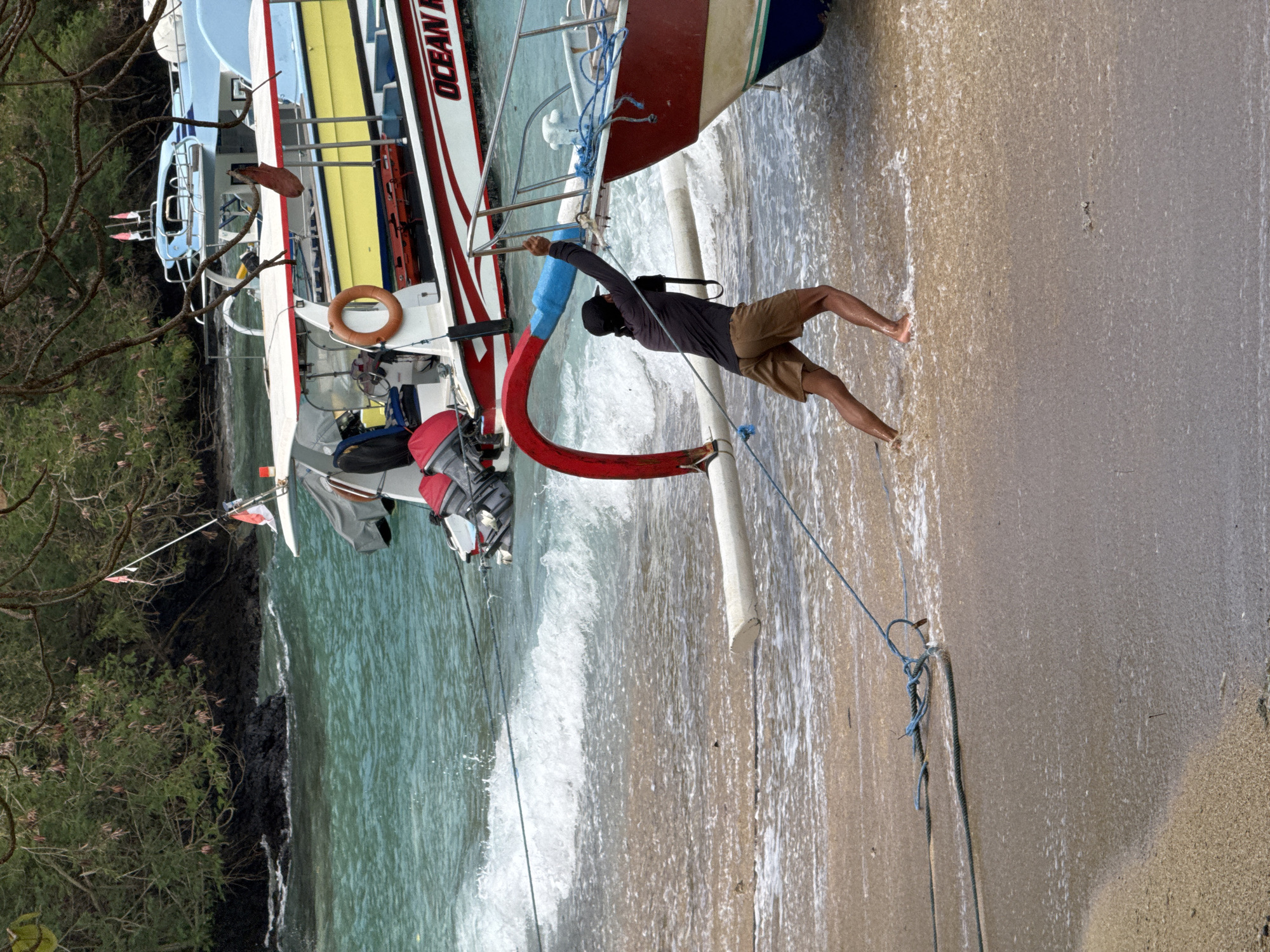 A person wearing a dark long-sleeved shirt and tan shorts is pulling a rope attached to a boat on a sandy beach in Manggis, Indonesia. The boat, which is red and white with blue trim, is partially in the water. Waves are washing onto the shore. In the background, other boats are docked. The environment is a coastal area with vegetation and rocky outcroppings visible in the distance.