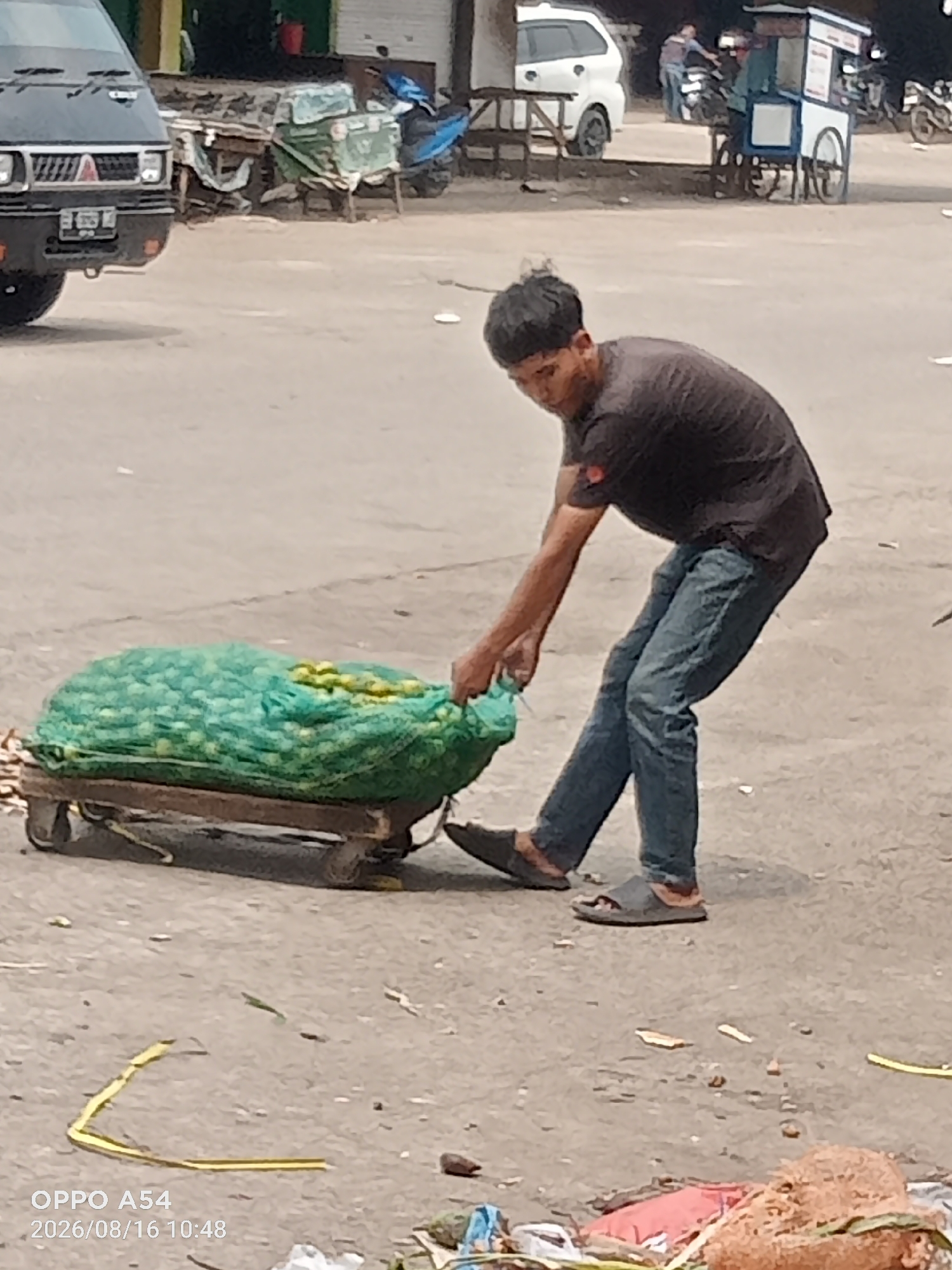 A man is depicted pulling a cart loaded with a large green net bag filled with yellow produce, likely fruit, in what appears to be an outdoor market setting in Jambi City, Indonesia. The man, wearing a dark t-shirt and blue jeans, is actively engaged in moving the cart, suggesting he is a vendor or a worker transporting goods. The cart itself is simple, with wooden planks and two visible wheels. In the background, a black van is parked to the left, and other market stalls or shops are visible, along with a white SUV and a food cart. The ground is paved and appears dusty, indicative of an urban environment. The overall scene captures a moment of daily commerce and labor.