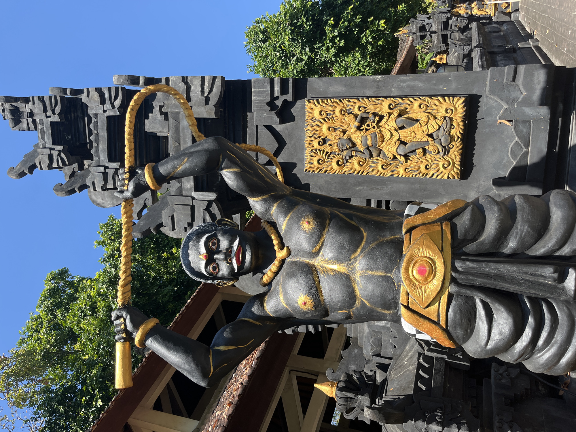 A statue of a muscular, dark-skinned figure with gold accents stands in front of a black stone temple structure under a clear blue sky. The statue has a red dot between its eyebrows, a white mustache, and is wearing a gold necklace, gold bracelets, and a gold belt with a red gem. It holds a thick, golden rope above its head. To the right of the statue, on the temple structure, is a gilded bas-relief of a multi-armed figure. The background shows a brown-tiled roof and lush green trees. This scene is located in Kuta Selatan, Indonesia.