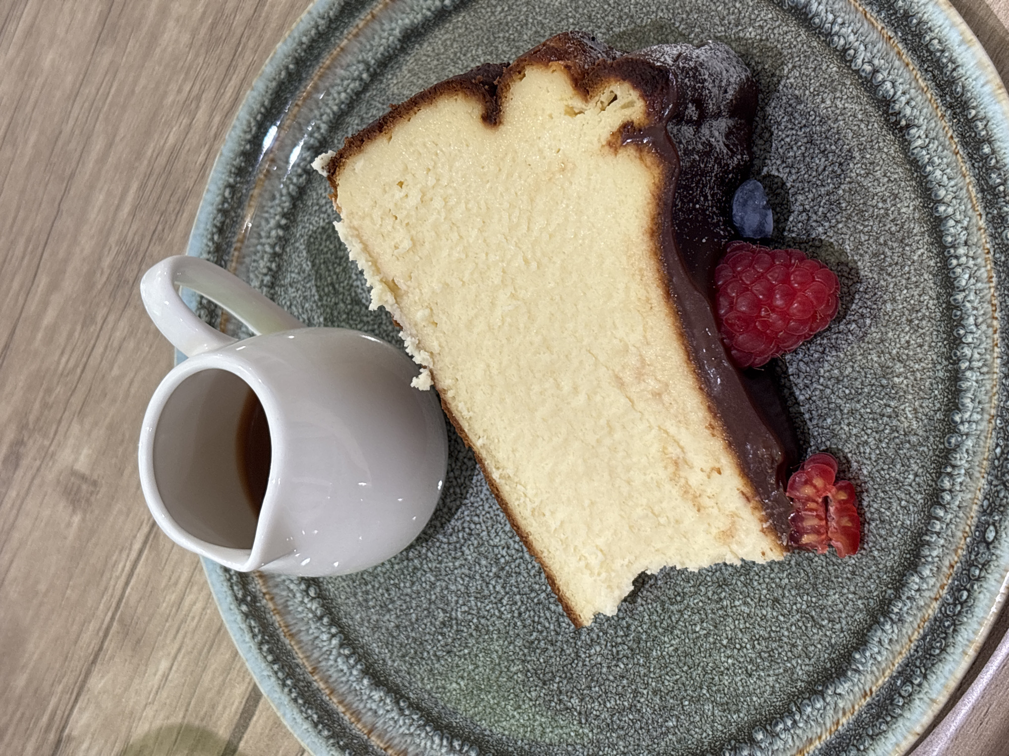 On a light-colored wooden surface is a round plate. The plate is a speckled grey-green color with a blue-grey trim. A slice of cheesecake with a chocolate topping is on the plate, garnished with raspberries and a blueberry. A small white pitcher, partially filled with brown liquid, sits to the left of the cheesecake slice. The image was taken from an overhead perspective. Location: Unknown, Unknown.
