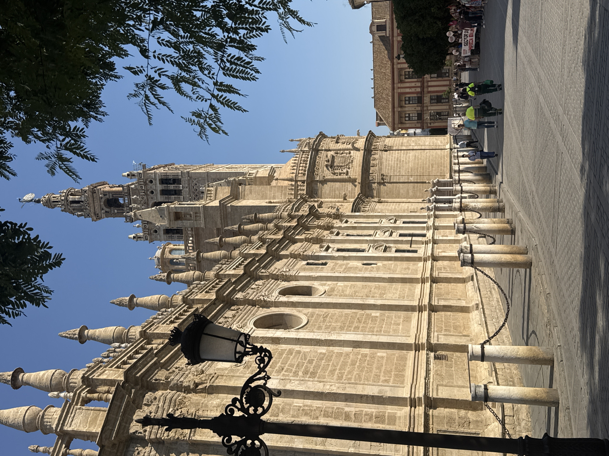 This media file depicts a sunny day in Sevilla, Spain, with a focus on the exterior of a large, ornate cathedral. The camera angle is angled upwards, emphasizing the grandeur of the architecture and the clear blue sky. Lush green foliage from a tree drapes into the top left corner of the frame, providing a natural contrast to the stone structure.
The cathedral itself is constructed from light-colored stone, showcasing intricate carvings and decorative elements. Multiple spires and architectural embellishments are visible, as is a large bell tower in the background. In the foreground, a decorative black lamppost stands prominently. Along the side of the cathedral, a row of thick, stone columns connected by chains line a paved walkway.
In the background, a group of people are gathered on the walkway, some holding signs. The signs bear text, though it is not clearly legible in the provided image. The people appear to be participating in a public gathering or demonstration. The time of day is indicated by the bright sunlight casting distinct shadows on the pavement and the building, suggesting it is mid-morning or early afternoon. The weather is clearly fair and sunny. The presence of the specific architectural style and the mention of Sevilla, Spain, place the scene in a historical and cultural context known for its impressive cathedrals.