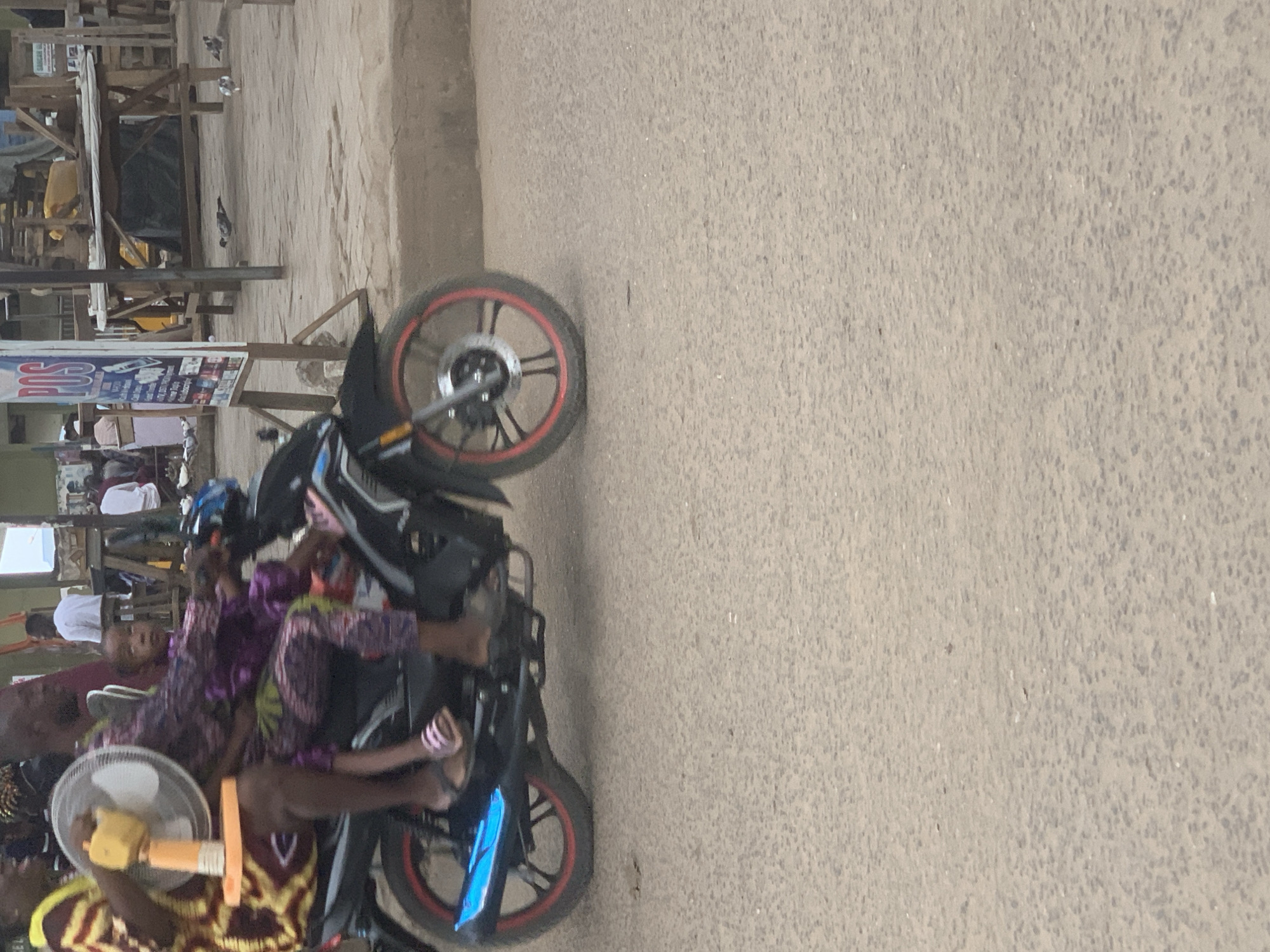 In Shagamu, Nigeria, a man and two children are riding on a motorcycle on a paved road. The man is in the front, operating the motorcycle. A younger child is seated between the man and an adult woman who is riding behind the man. The woman is holding a portable fan. The motorcycle has a blue exhaust pipe. In the background, there are shops and a sign that reads "POS". Several pigeons are on the ground in the background.