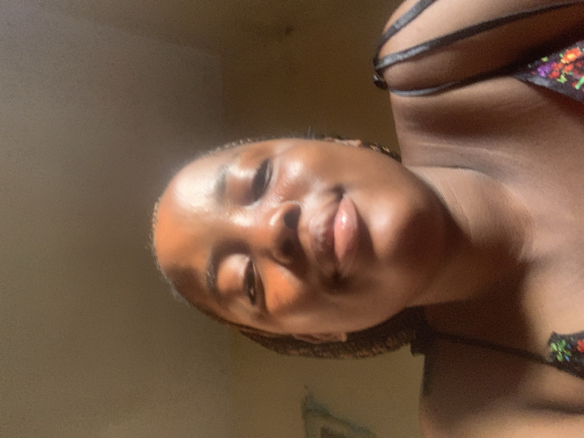 A close-up, low-angle selfie captures a person with dark skin, oriented horizontally on their left side, looking directly at the camera. The individual, likely female, has strong highlights on their forehead, nose, and full, glossy lips, suggesting a radiant complexion under natural light. A slight smirk or gentle smile is visible. They are wearing a dark-colored top with thin straps, and a portion of a colorful, possibly floral or abstract patterned bra or top is visible on the right side of the frame. The background is a simple, warm-toned, plain surface, indicative of an indoor setting. The lighting appears to be natural, casting bright highlights on the skin and suggesting a daytime environment. The image is slightly grainy, consistent with a mobile phone photograph. No text is visible. This scene is contextualized to Umuode, Nigeria.