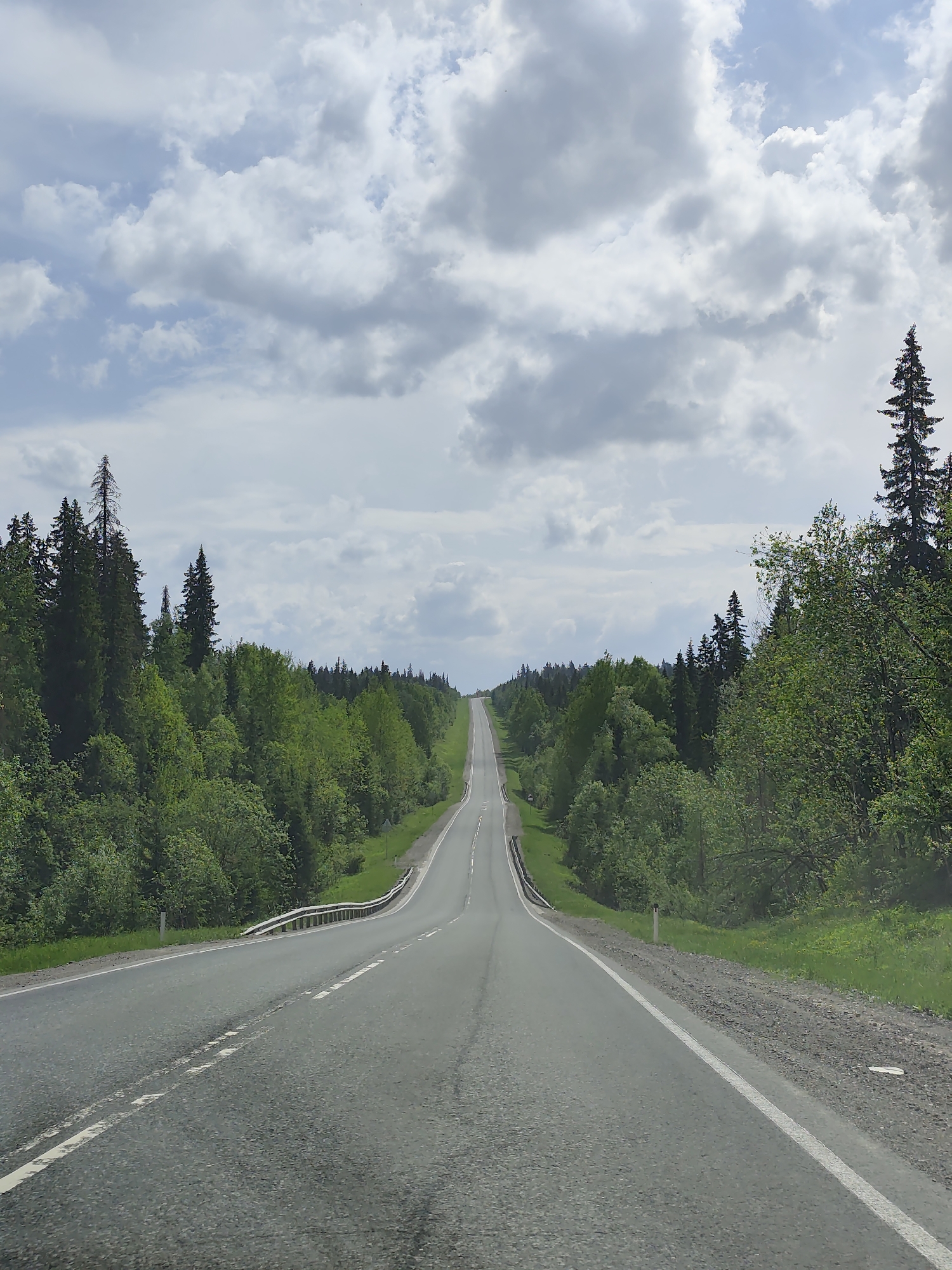 A long, straight asphalt road stretches into the distance, flanked by green forests on either side, under a cloudy sky. The road has white lane markings. The location is Gremyachinsk, Russia. There are no people visible.