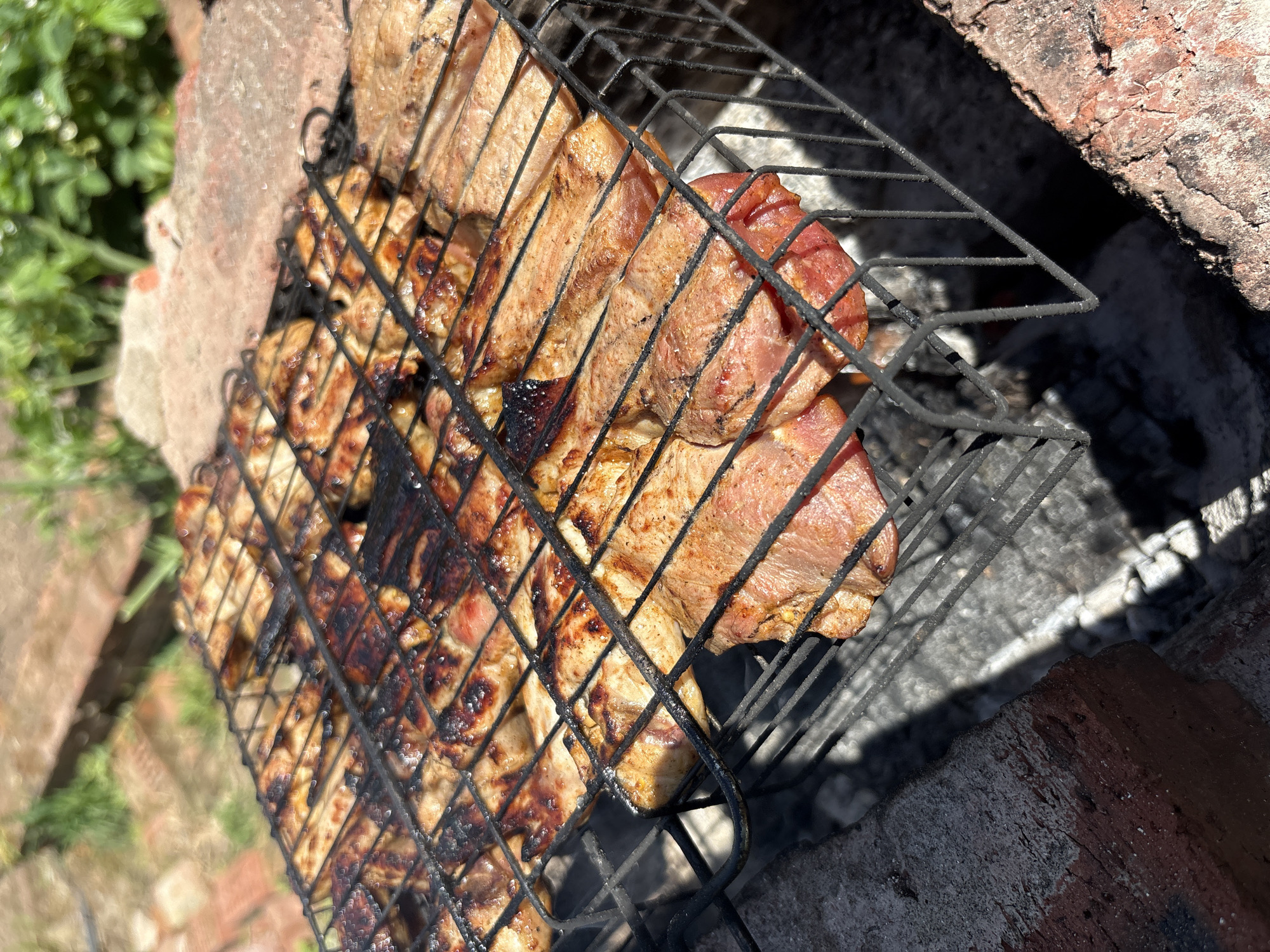 This image captures a close-up, elevated view of an outdoor grilling scene in Italmas, Russia. Various cuts of meat, appearing to be chicken pieces and possibly some pork chops, are cooking on a rectangular metal grill grate. The meat on the left side shows distinct char marks and a well-browned, caramelized surface, indicating it is nearing completion or has been cooking for some time. The meat on the right exhibits lighter browning, with some pieces still showing a pinker, less cooked appearance.
The grill itself is a black, wire-mesh design, likely a hinged basket-style grate, holding the food directly over a bed of glowing coals and white ash, partially visible beneath the grate. The grilling apparatus appears to be constructed from rough, reddish-brown bricks, forming an open-air barbecue pit.
The scene is brightly lit by direct sunlight, casting sharp, elongated shadows from the grill and food onto the ash and the inner walls of the brick structure. The background is out of focus, revealing hints of a paved area or more bricks on the left, and blurred green foliage on the right, suggesting a garden or yard environment. No individuals are visible in the frame.
