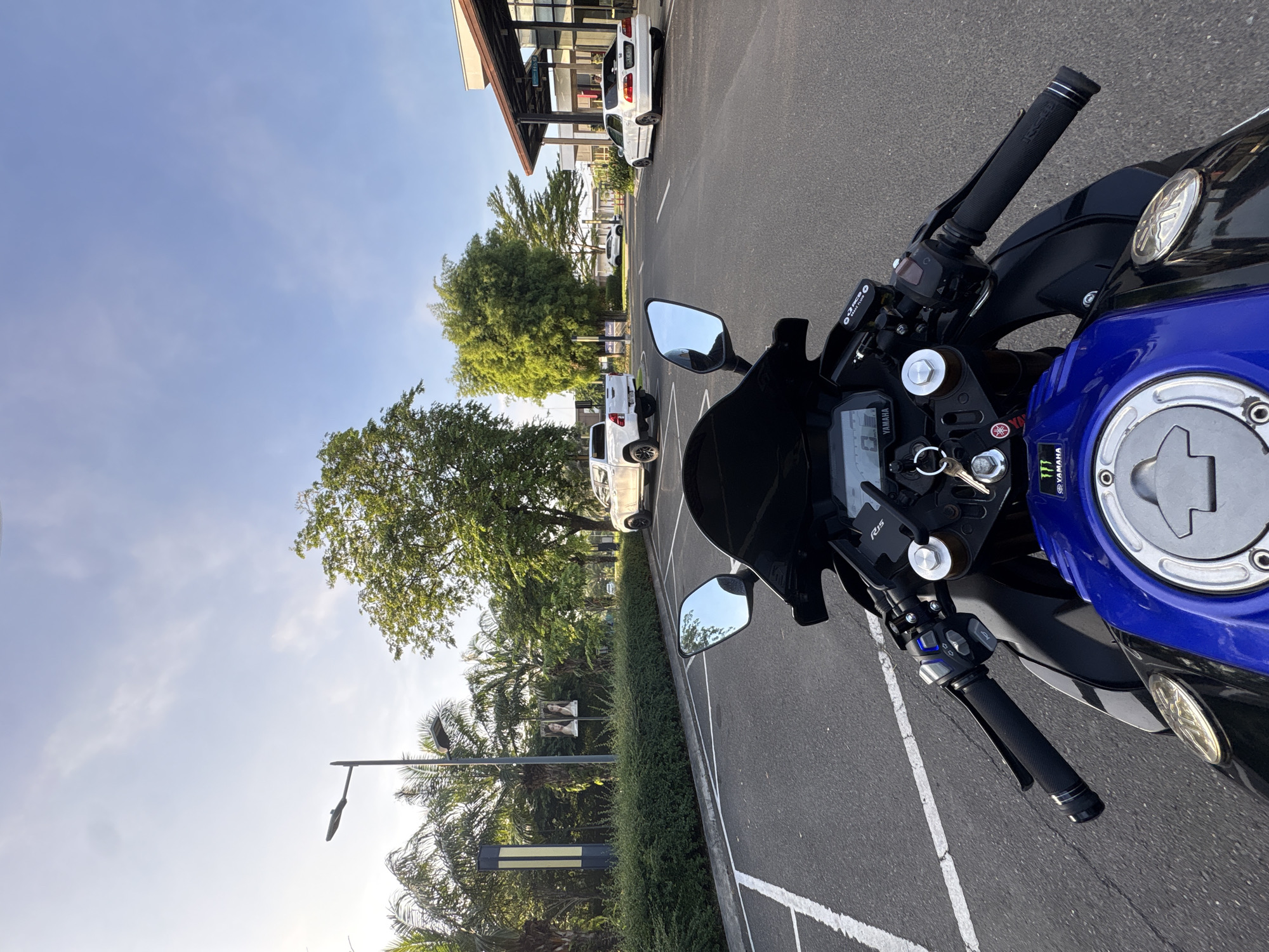 The image is taken from the perspective of someone sitting on a blue and black motorcycle in a parking lot. The motorcycle is the primary subject in the foreground, with its handlebars, dashboard, and fuel tank visible. In the background, there is a paved road with white parking lines, a white pickup truck, a white sedan, and a building with a modern facade. Lush green trees and palm trees are also visible, suggesting a tropical environment. The sky is clear with a few wispy clouds. The scene appears to be set in a well-maintained commercial or residential area. Based on the presence of palm trees, the clear blue sky, and the overall ambiance, the location is consistent with a place like Davao City, Philippines.