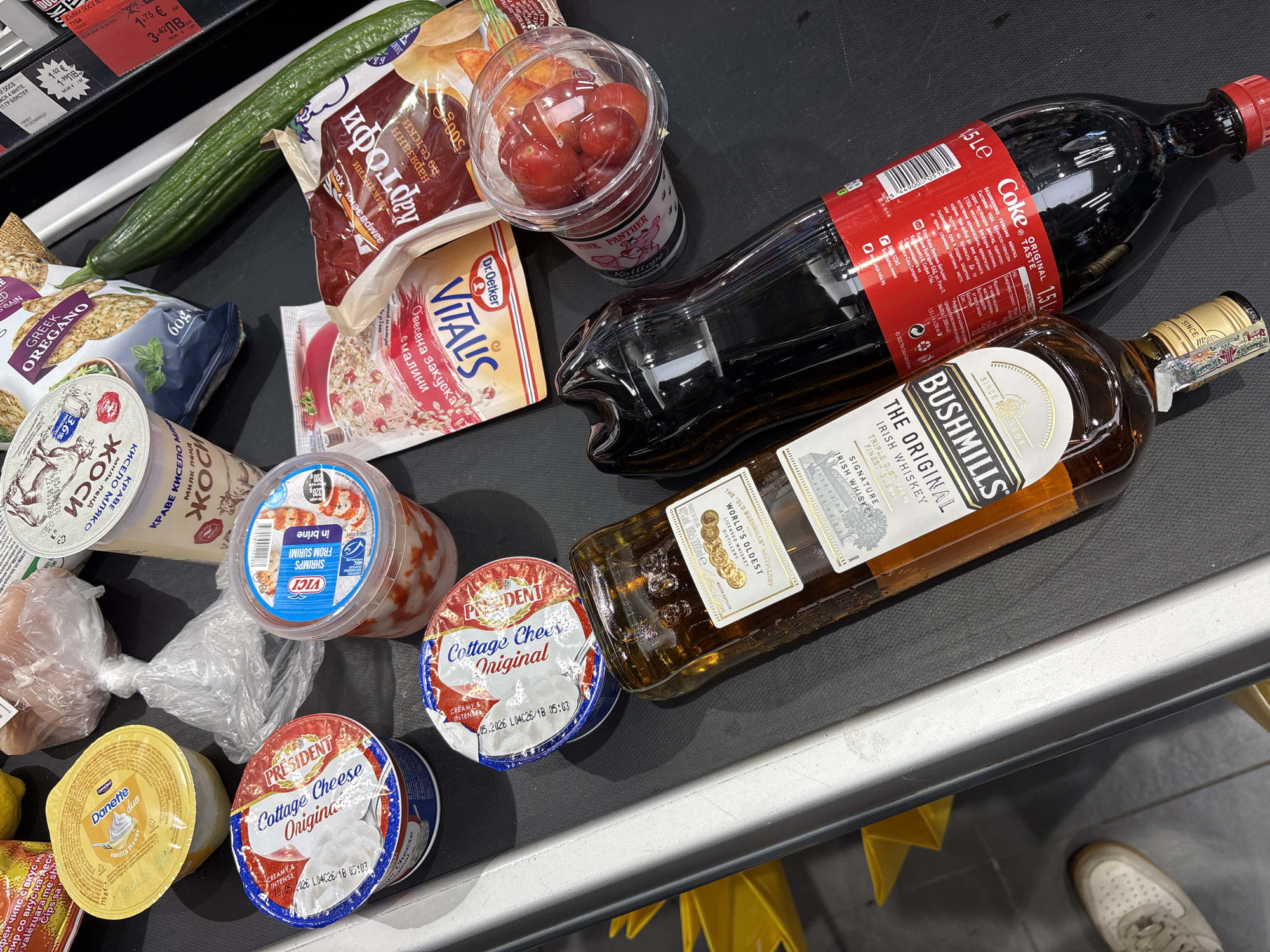 The image displays a collection of groceries on a black supermarket conveyor belt, indicating an ongoing checkout process. The setting is an indoor grocery store environment. Part of a person's foot, clad in a white and grey sneaker, is visible on the bottom right, suggesting their presence near the checkout.
A variety of items are present: beverages include a large 1.5-liter bottle of Coca-Cola Original Taste and a 1-liter bottle of Bushmills The Original Irish Whiskey. Dairy products feature two tubs of President Cottage Cheese Original, a yellow Danette Duo vanilla-flavored dessert cup, and two cups of Milk Land dairy products, one labeled "КРАВЕ КИСЕЛО МЛЯКО" (Cow's Sour Milk).
Fresh produce includes a green cucumber and a clear plastic cup of cherry tomatoes, branded "PINK PANTHER Kalliopo." Packaged goods consist of a bag of Greek Oregano flavored chips/crackers, a bag of frozen rustic-cut potatoes ("КАРТОФИ нарязан по селски"), a sachet of Dr. Oetker VITALIS Oatmeal breakfast with raspberries ("Овесена закуска с малини"), a container of VICI Shrimps from Surimi in brine, a small plastic bag likely containing raw meat, and a bag of Fen Chips with a meat flavor ("Фен чипс с вкус на месо").
Several texts provide contextual clues: the Coca-Cola label explicitly mentions "Кока-Кола Хеленик Ботълинг България ЕАД, София 1766." Price tags visible in the background show values in both Euro (€) and Bulgarian Lev (ЛВ), confirming the location as Sofia, Bulgaria. Other visible text includes brand names, product descriptions, and expiry dates like "05.2026." No specific time of day or emotional states are discernible.