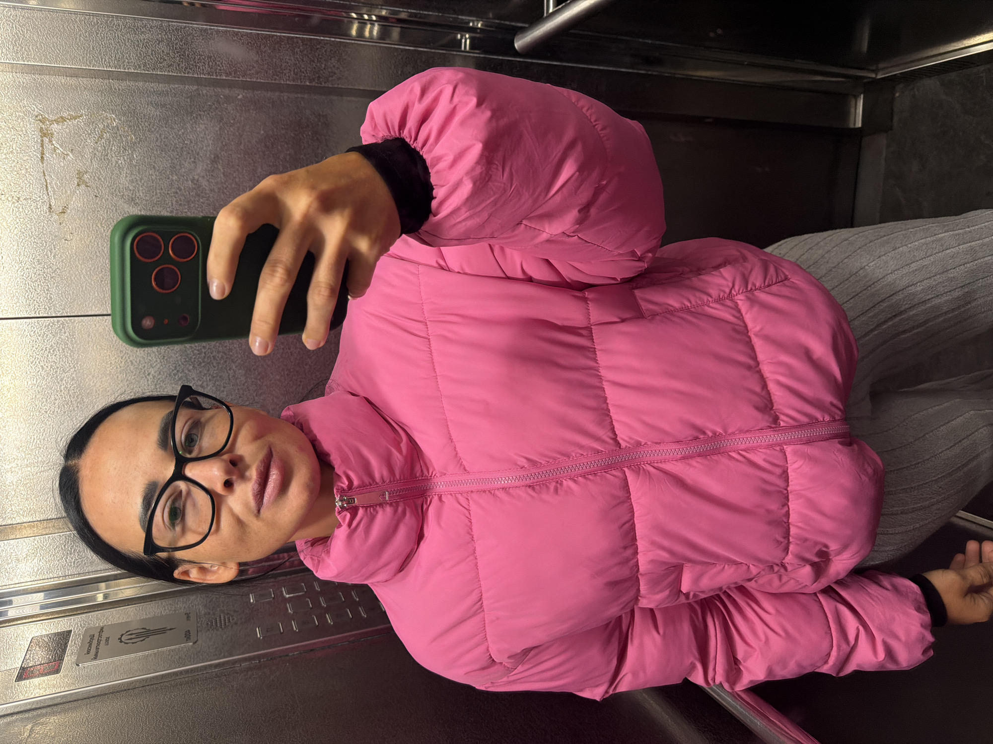 A woman wearing glasses and a pink puffer jacket takes a selfie with a phone in an elevator. The interior of the elevator is silver, with visible metal bars. An information panel is present. There is no indication of the exact location in the city of Saint Petersburg, Russia.
