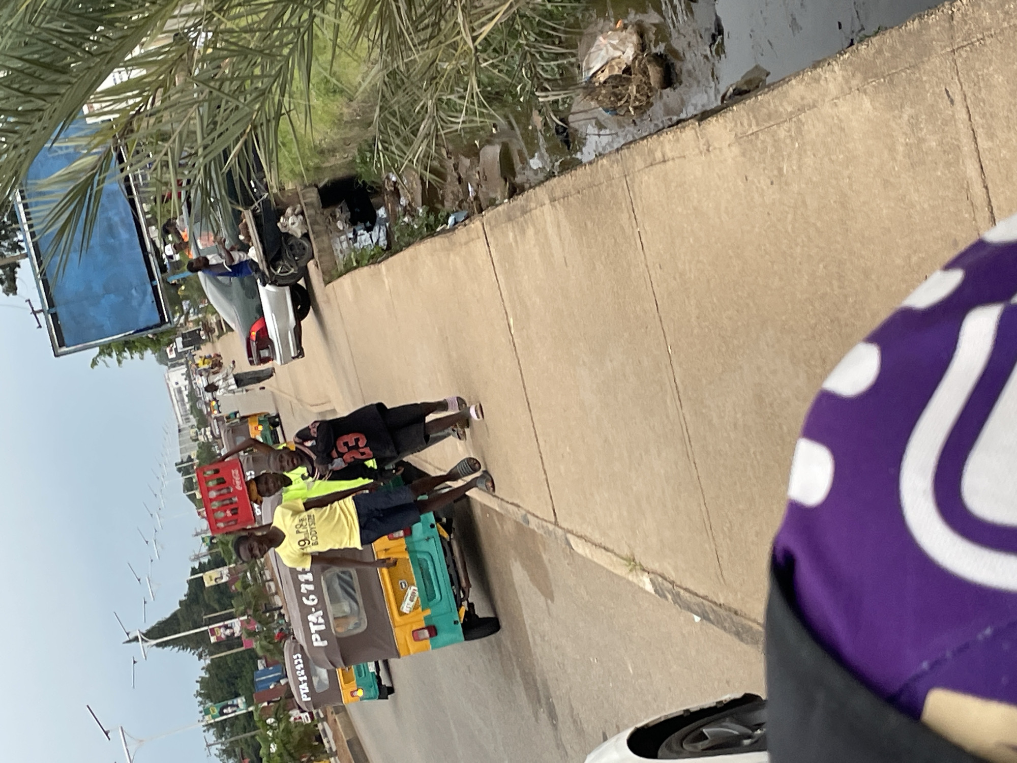In Barakin Lerawi, Nigeria, a group of young men walk along a sidewalk next to a busy road. One man in a yellow t-shirt carries a red crate of Coca-Cola bottles on his head. Behind him, another young man in a neon green t-shirt and one in a black jersey with the number 23 on it walk beside him. In the background, several auto-rickshaws, a silver sedan, and other vehicles are visible on the road. A drainage ditch filled with debris runs alongside the sidewalk, and palm fronds overhang from a tree on the right side of the image. The sky is overcast.