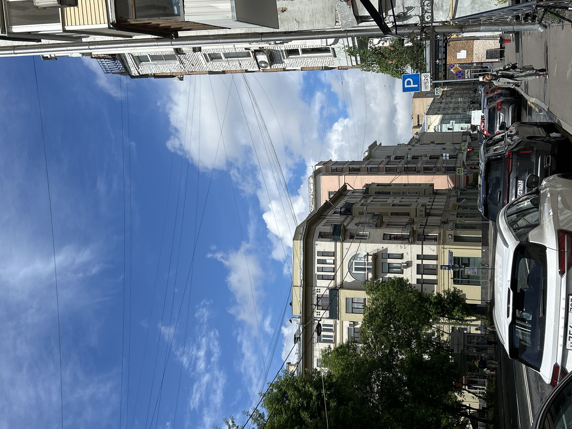 The image depicts a street scene in Unknown, Unknown. Buildings line the street on either side, with power lines running overhead against a backdrop of blue sky and scattered clouds. Cars are parked along the curb, including a white car with a visible license plate that ends in "797." A pedestrian is visible on the sidewalk, and a parking sign is present. The buildings are a mix of architectural styles and colors, with one building notably painted pink. Trees are interspersed among the buildings and along the street.