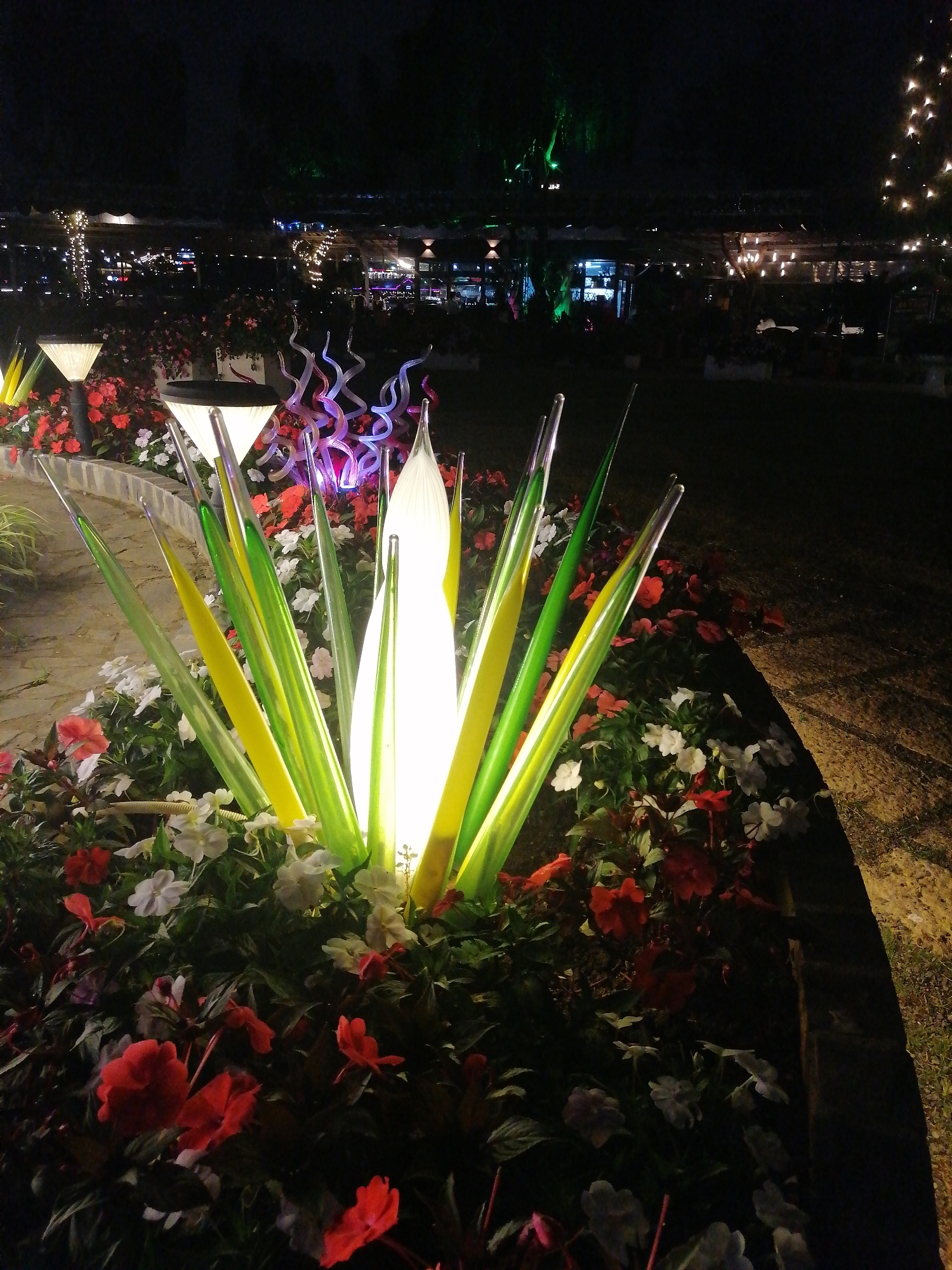 This nighttime photograph captures a vibrantly illuminated garden scene, likely in Da Lat, Vietnam. The foreground is dominated by a striking art installation resembling an agave plant, featuring a tall, central white light emanating from a translucent core, surrounded by numerous sharp, glowing spikes of green and yellow glass or acrylic. This centerpiece is nestled within a lush flowerbed filled with small red and white blossoms.
To the left of the main sculpture, two modern, triangular solar path lights cast a warm glow, while slightly behind and to the right, another abstract light sculpture with swirling, translucent purple and pink elements adds to the eclectic display. The ground nearby appears to be a textured path.
In the mid-ground and background, numerous small, warm string lights are draped across trees and structures, creating a festive and inviting ambiance. Some trees in the deeper background are subtly illuminated with green light. The dark night sky emphasizes the artificial illumination, creating a magical atmosphere. No people or visible text are present, suggesting a tranquil moment in a beautifully designed public garden or attraction.
