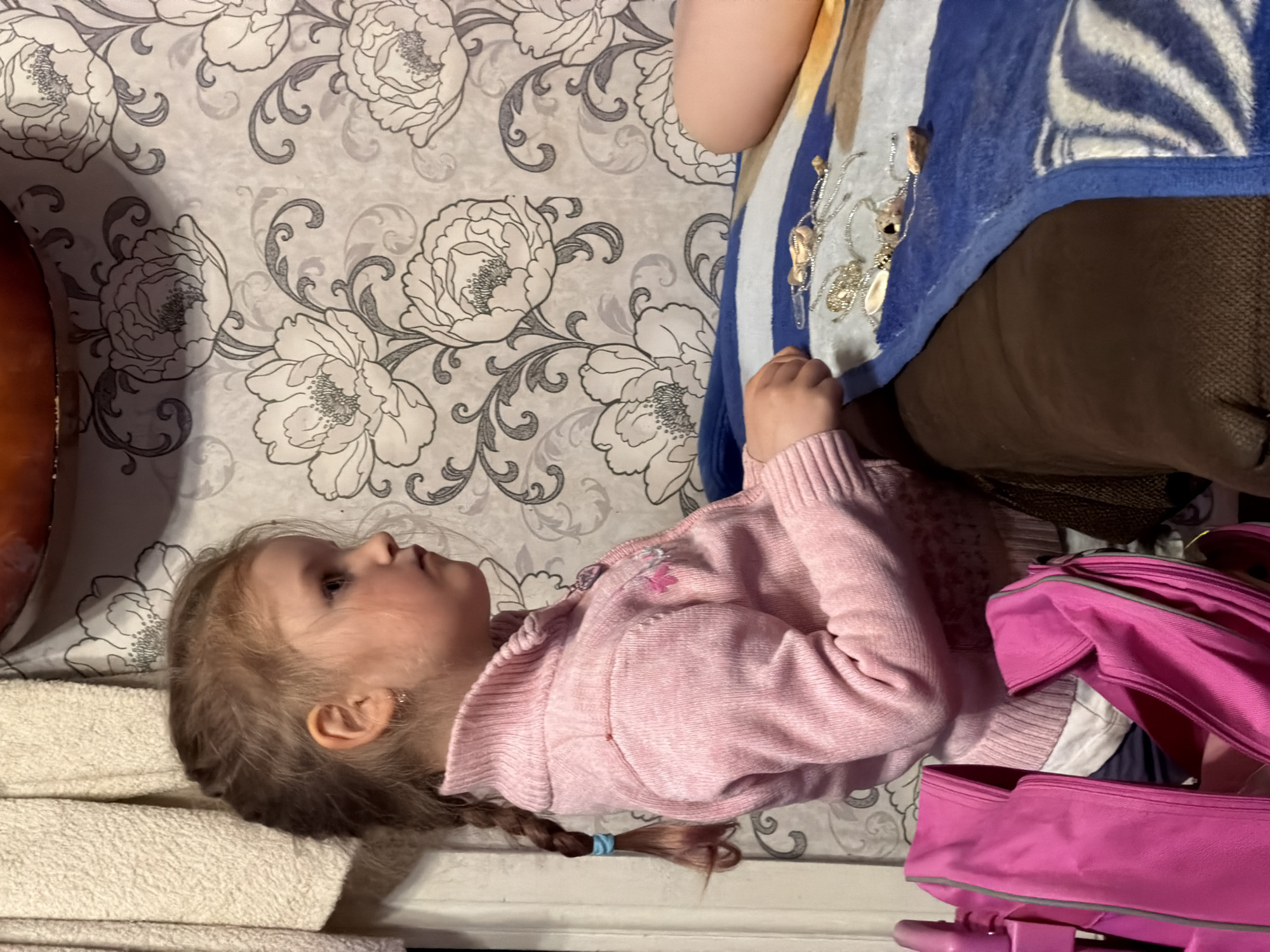 This image, rotated 90 degrees counter-clockwise, captures a young girl lying or reclining indoors. The setting appears to be a home in Заболотово, Russia. The girl, likely a toddler or preschooler, has light brown hair styled in two braids, one of which is visible with a light blue elastic band. She is wearing a light pink zippered cardigan, possibly over a white top, and a small gold earring is visible in her right ear. Her gaze is directed upwards and forward, displaying a neutral, thoughtful, or observant expression.
The background wall features light-colored wallpaper with a repeating pattern of large white and gray floral designs, accented by dark gray scrollwork. To the left of the girl's head, a stack of white, textured towels or fabric is visible. In the upper right portion of the image, part of another person's arm is seen, alongside a blue and white patterned blanket or throw that features decorative, seashell-like ornaments sewn onto its surface. This blanket covers a brown, textured surface, likely a sofa or chair.
To the girl's right, a bright pink backpack with gray trim is prominent, appearing to be partially open. Below it, another pink item, possibly a rolling suitcase handle or a second bag, is visible. The lighting is soft and ambient, characteristic of an indoor setting. There is no visible text in the image. The overall scene conveys a quiet moment of repose or observation.