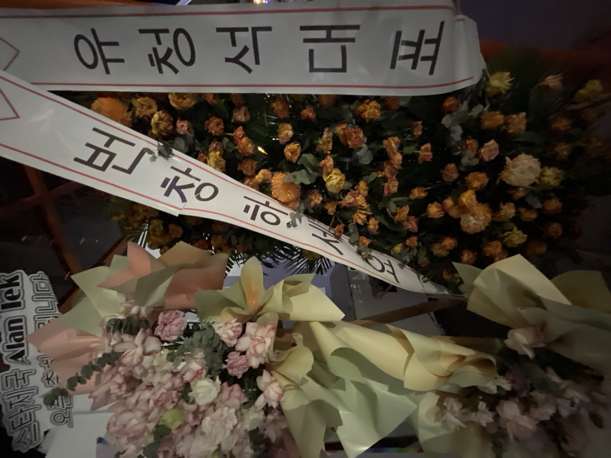The image shows a floral arrangement with a white ribbon bearing Korean text. The ribbon is diagonally placed over the flowers. On the right side of the ribbon, the text reads "유청석대". On the left side of the ribbon, the text reads "번창하". The arrangement itself consists of pink and white flowers on the left and orange flowers on the right, with some greenery and decorative paper. The background is dark and out of focus, suggesting it might be nighttime or an indoor setting with low lighting. No specific location in Da Nang, Vietnam, is identifiable from the image.