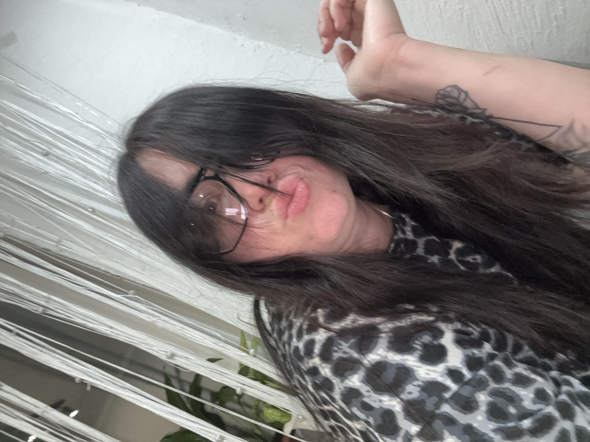 An overhead, tilted indoor shot captures a person with long, dark hair covering part of their face, posing with a pout. The person wears dark-rimmed glasses and a black, white, and gray leopard print top. Their right arm is visible in the upper right corner, bent at the elbow, revealing a black outline tattoo on the inner forearm, resembling a stylized rose or flower.
The background features vertical strands of white material, possibly string curtains or blinds, creating a soft, textured blur. Hints of green foliage, likely from a houseplant, are faintly visible behind these strands in the lower left quadrant. The overall lighting is soft and diffused, characteristic of an indoor environment. The facial expression conveys a playful or perhaps slightly sassy mood. No discernible text is visible in the image.
