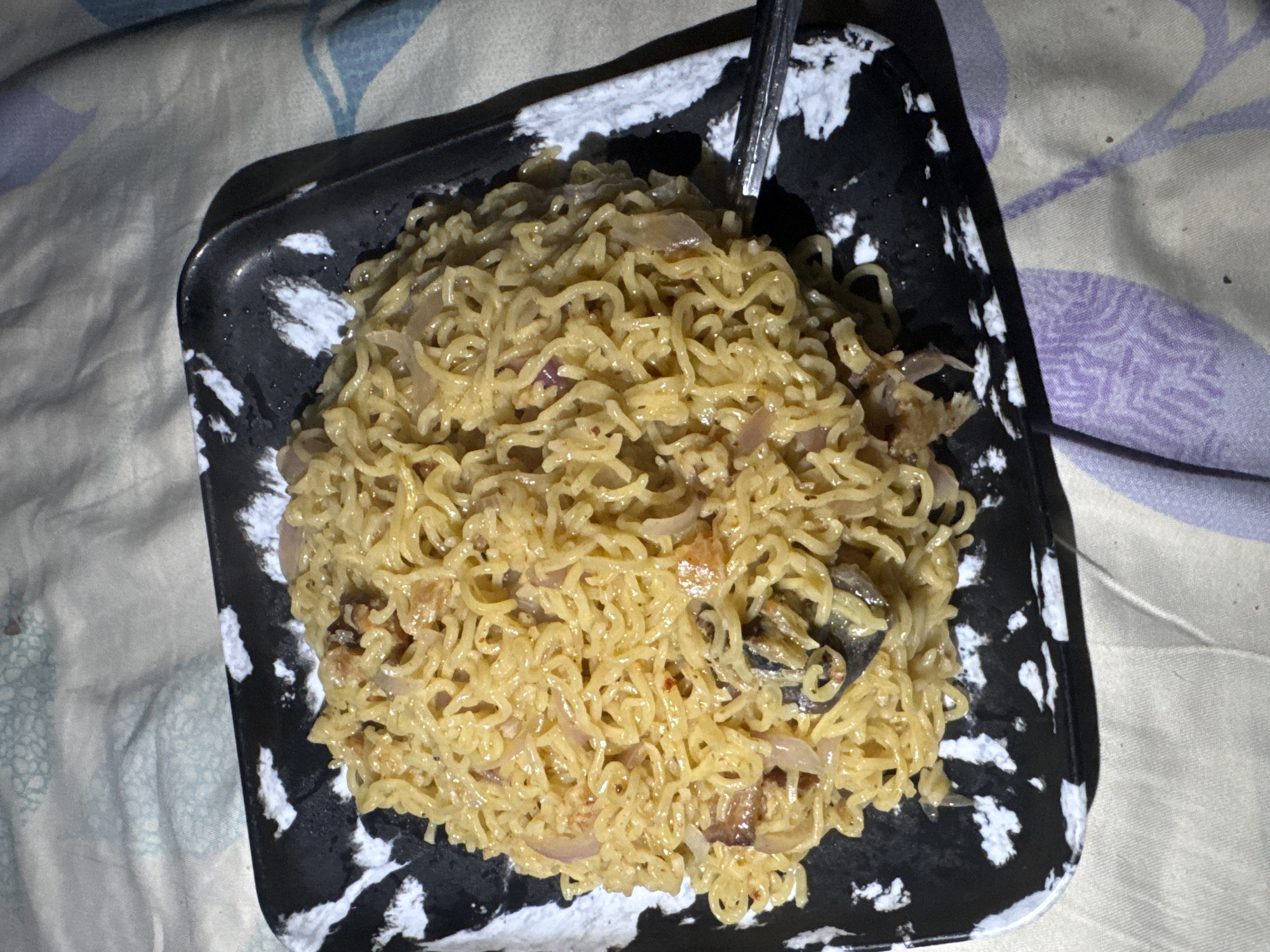 An overhead shot, taken in Ihiagwa, Nigeria, displays a square black plate filled with cooked instant noodles. The plate features a unique design with irregular white splatters across its dark surface. A metal spoon is partially visible, inserted into the noodles on the right side of the plate, indicating the meal is ready for consumption or is being eaten.
The noodles are light yellowish-brown, curly, and appear well-cooked, mixed with translucent, light brown slices of what seem to be sautéed red onions, and possibly other small, darker solid ingredients. The noodles are coated in a glossy sauce or seasoning.
The plate is set on a patterned fabric surface, likely a bedsheet, which serves as the background. On the left side of the frame, the fabric shows abstract patterns in light blue and white, while the right side features similar abstract patterns in purple and white. The overall lighting is dim and warm, suggesting an indoor setting during the evening or nighttime. No individuals are present in the image, and no text is visible.