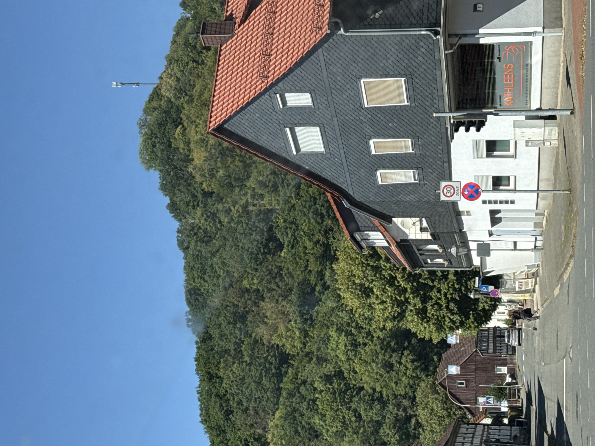 The image captures a street scene in Bad Lauterberg im Harz, Germany, with a dense green forest covering a hillside in the background. A cell tower is visible on top of the hill. In the foreground, a dark-colored, modern house with a red tiled roof is prominently featured on the right. Its facade is covered in dark grey slate tiles. Next to it, a building displays a sign for "KATHLEENS FAHRSCHULE" (Kathleen's Driving School). On the left side of the street, traditional half-timbered houses line the road. A silver car is driving away from the camera down the street. A person is seen walking across the street, pushing a broom. Traffic signs, including a 30 km/h speed limit sign and a no parking sign, are visible. The sky is clear and blue.