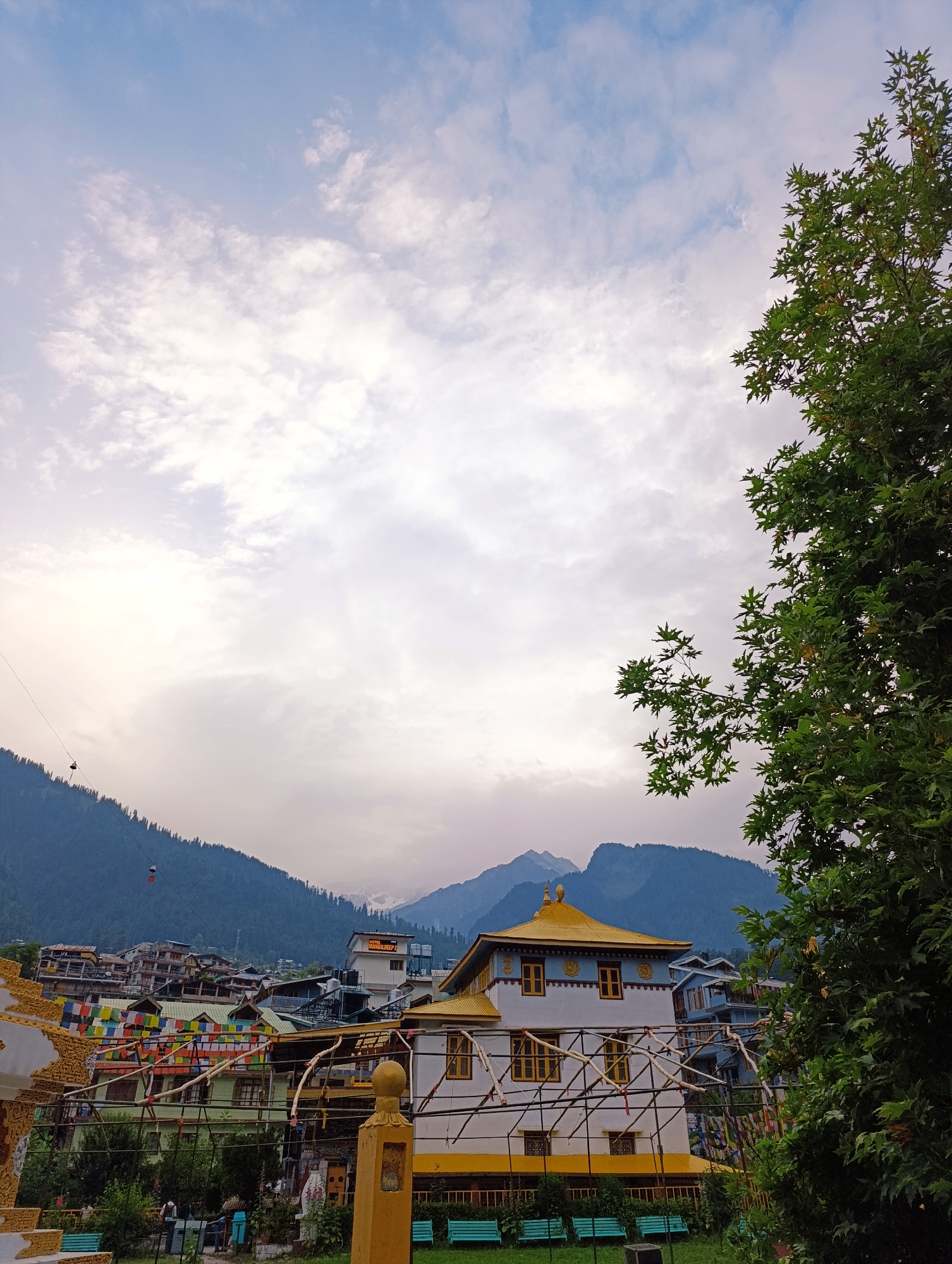 The image depicts a view of the Kyidomang monastery in Manali, India, nestled amidst the Himalayas. The monastery's white facade is adorned with a striking golden roof and yellow-trimmed windows, reflecting the distinct architectural style of the region. In the foreground, a lush green tree with vibrant foliage partially obscures the view of the monastery. Beyond the monastery, the city of Manali unfolds, characterized by its densely packed buildings and a backdrop of imposing, mist-shrouded mountains. Prayer flags, a common sight in Buddhist regions, are strung across the buildings, adding a splash of color to the scene. A cable car system is visible in the distance, traversing the mountainous terrain. The sky above is a canvas of soft clouds, hinting at either an approaching storm or the remnants of one. A few individuals are present in the foreground, appearing to be engaged in casual activities. The overall atmosphere is one of serene beauty and cultural richness.