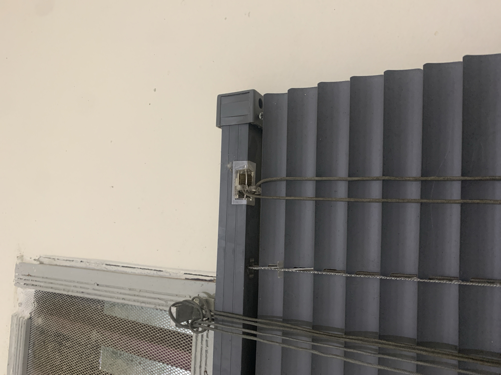 The image shows a close-up view of a grey horizontal blind. The blind is partially lowered, with several slats visible. Three grey cords hang from the top of the blind, the one on the left is tied in a knot. A clear plastic bracket is attached to the top of the blind mechanism, holding a cord in place. To the left of the blind, a portion of a window frame is visible, covered by a metal mesh screen. The wall behind the blind is a light beige color. The scene appears to be indoors, possibly in a residential or commercial setting. There are no people or animals present in the image. The location in Kuala Pembuang, Indonesia, is not discernible from the image itself.