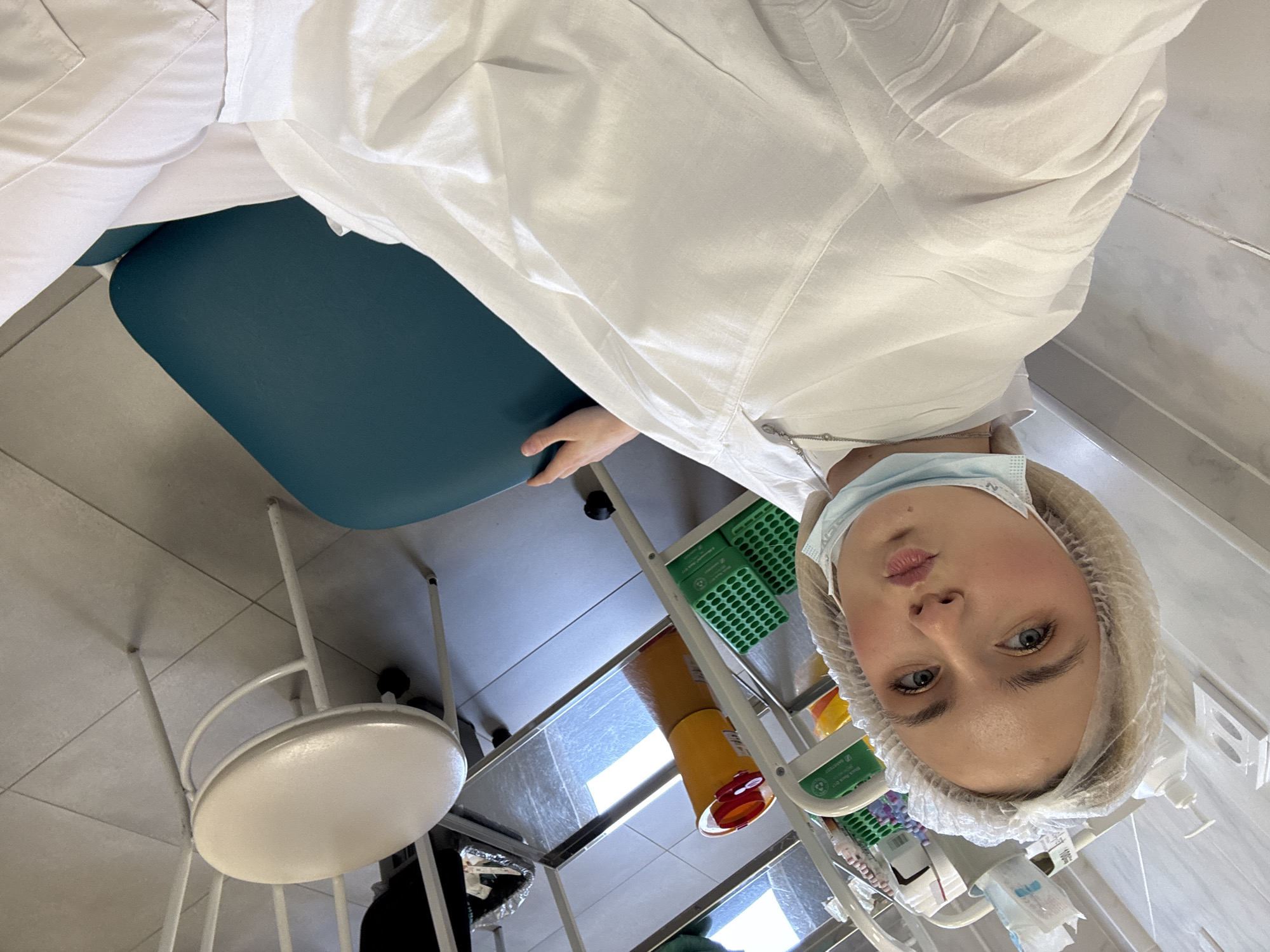 A person wearing a white medical coat, a surgical mask, and a disposable hair net is looking directly at the camera. The person's facial expression is neutral, with slightly pursed lips. The location appears to be inside a medical or dental office, evidenced by the equipment in the background. There's a dark teal chair to the person's left, a rolling cart with various medical supplies including test tube racks and waste containers, and a stool with a white seat. The flooring is tiled. There is no indication of the location within Saint Petersburg, Russia.
