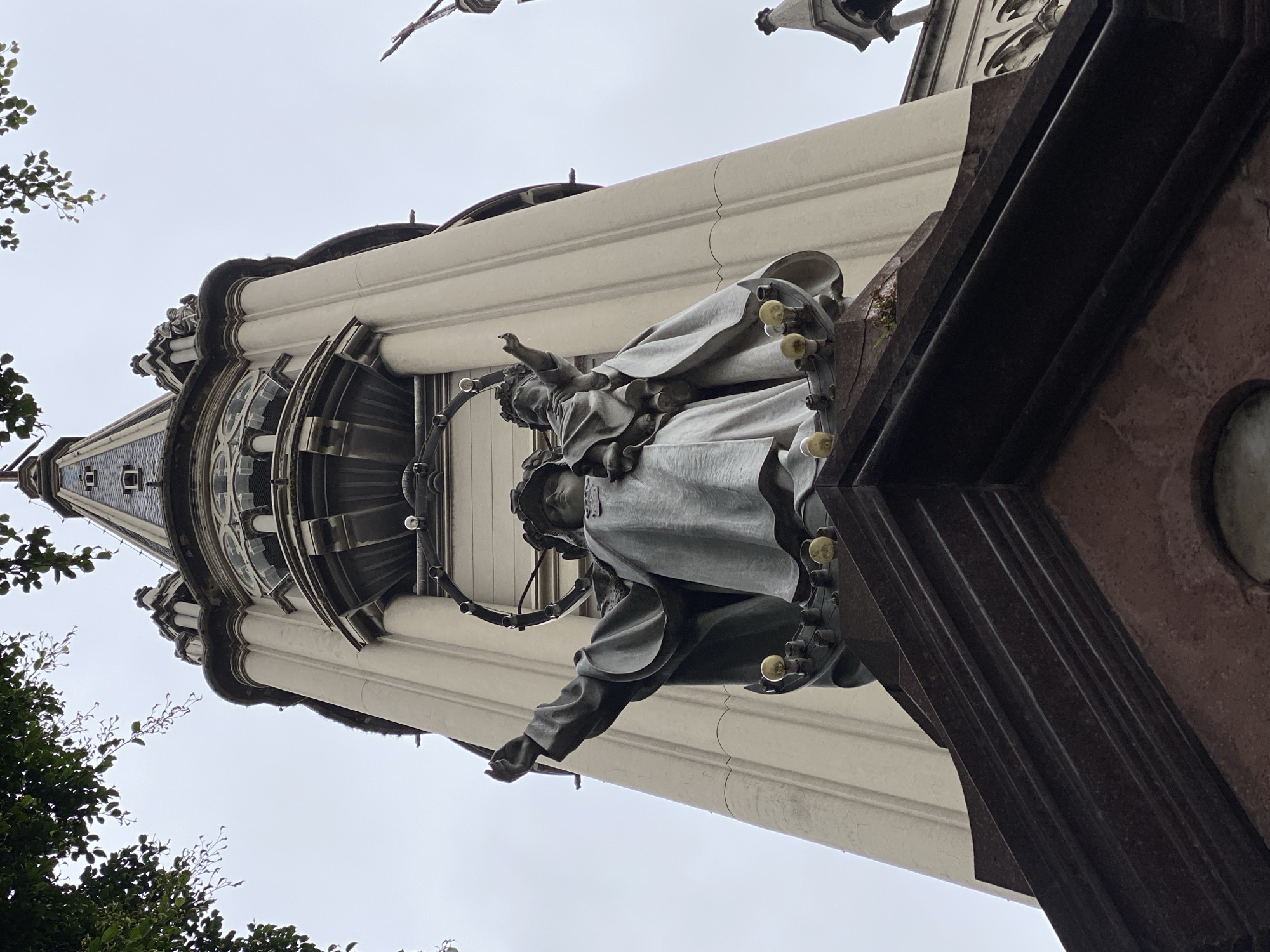The image shows a low-angle view of a building's architectural details. The building appears to be a tower or steeple with ornate decoration. A gray sculpture of a robed figure with another smaller figure is mounted on the building's facade. The structure is characterized by a combination of white, gray, and brown stone, with visible vertical fluting and molded details. The sky is overcast. Part of a tree is visible in the lower left. The building is located in Buenos Aires, Argentina.