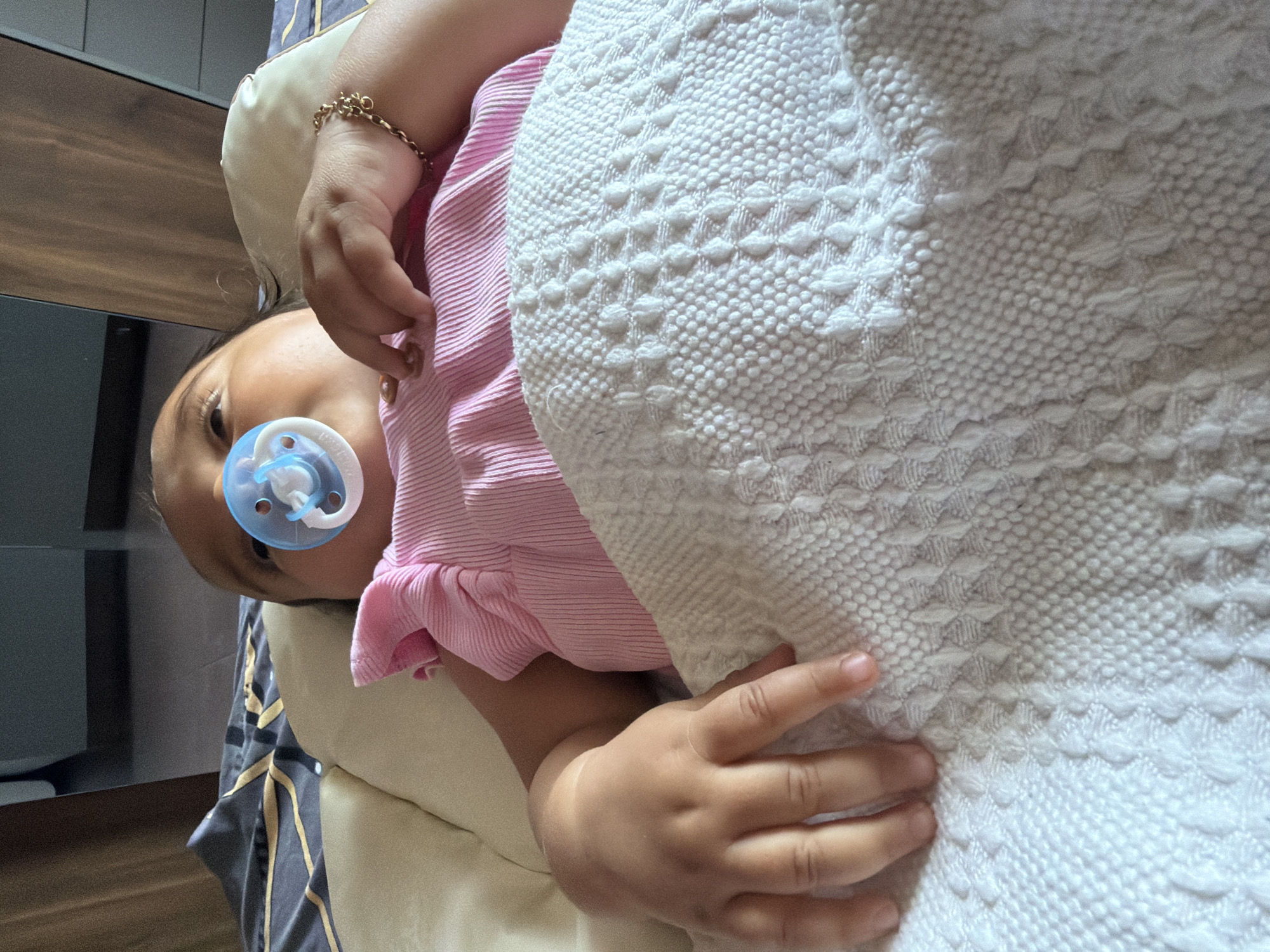 A baby is lying on a bed in Razhevo Konare, Bulgaria, with a pacifier in its mouth. The baby is wearing a pink shirt and is covered with a white blanket. The bed is in a room with wooden furniture.