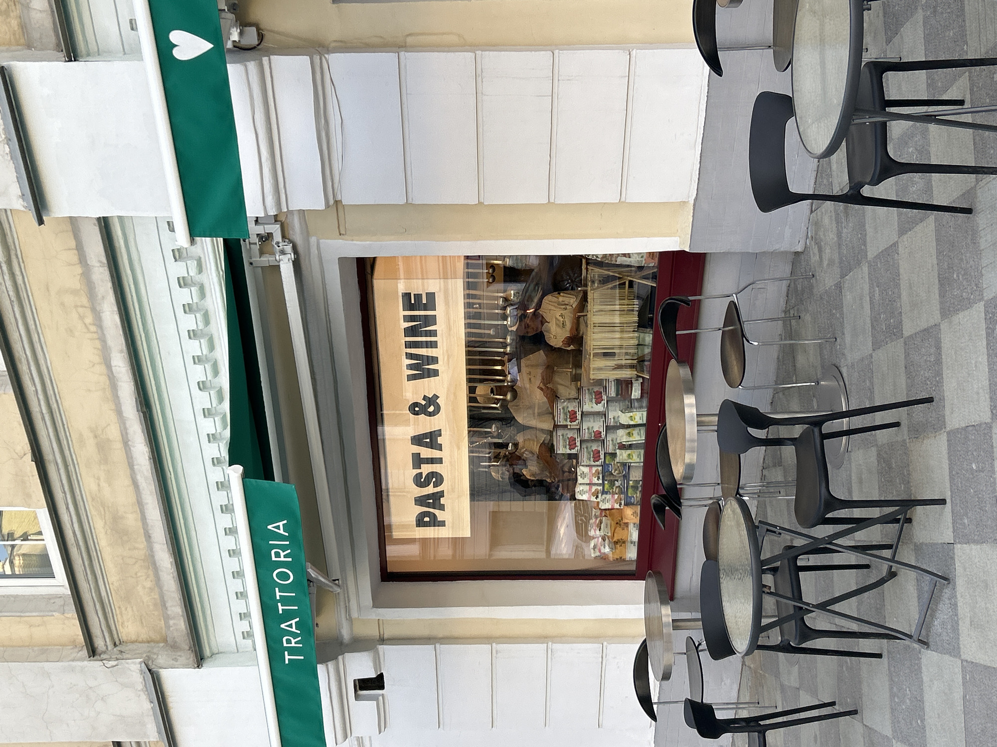 Here is a description of the image:
The image shows the exterior of a restaurant called "Trattoria" with the words "PASTA & WINE" displayed in the window. The restaurant has a green awning with a white heart. Tables and chairs are placed outside the restaurant on a checkered pavement. The city location is not specified, denoted as Unknown, Unknown.