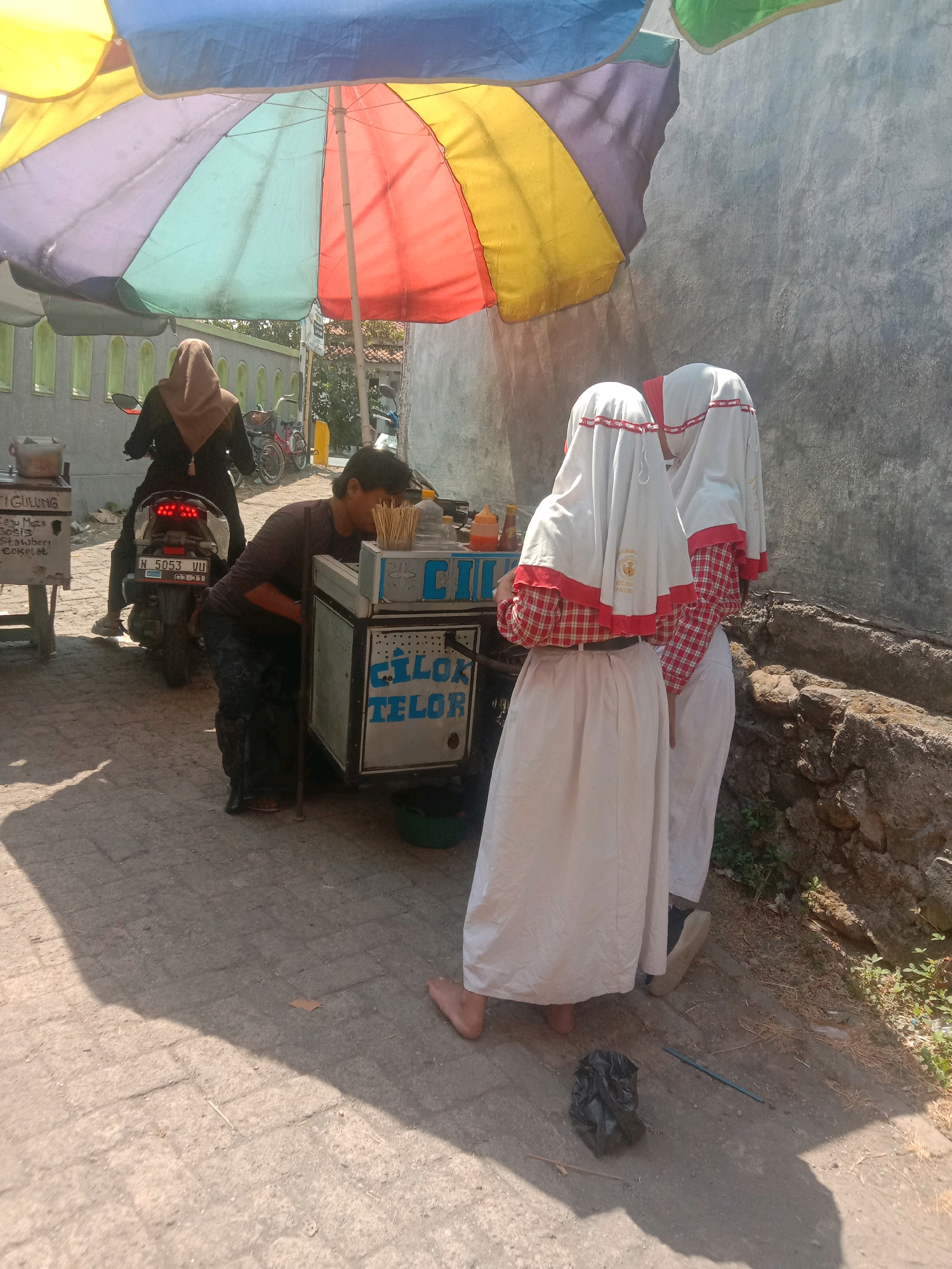 The image depicts an outdoor scene in Karang Anyar, Indonesia, under a large, colorful umbrella. Two young females wearing white hijabs and red and white checkered shirts are standing near a food cart. The food cart has text that appears to be in Indonesian, with the words "CILOK TELOR" prominently displayed. One person, likely a vendor, is attending to the cart. In the background, a person is seated on a motorcycle, and another motorcycle is parked nearby. The ground is paved with bricks. A wall with a textured surface is visible on the right side of the image, and some vegetation grows along its base. Sunlight casts shadows on the ground, indicating it is daytime. A black plastic bag and a blue stick-like object are on the ground in the foreground.