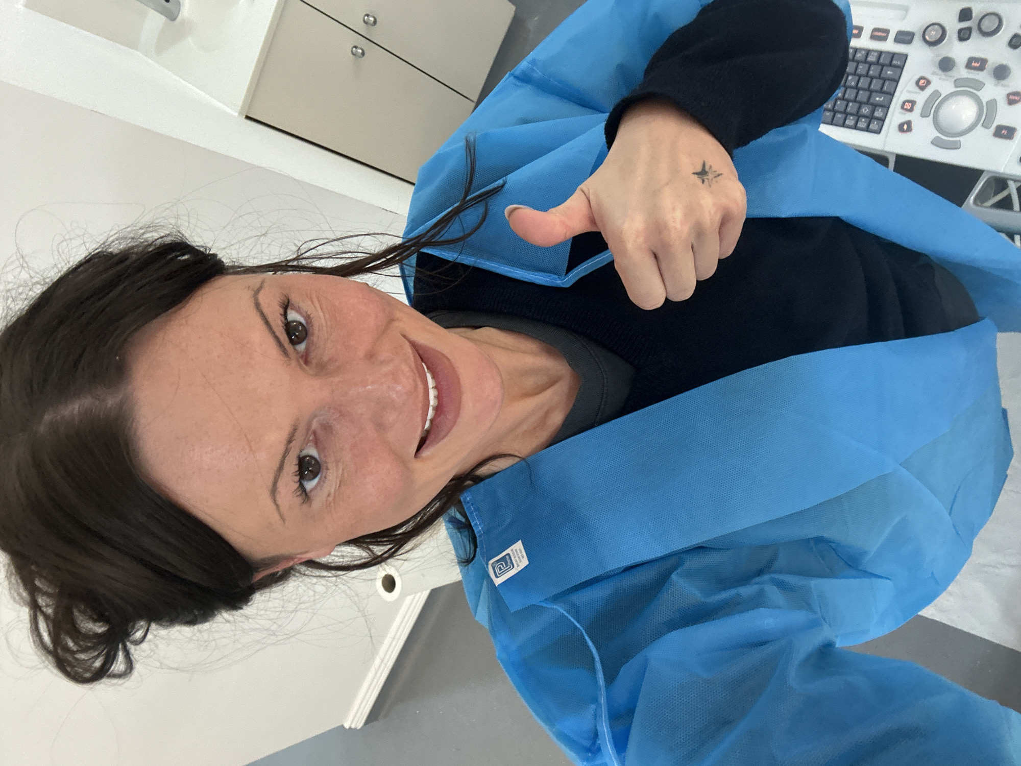 A woman with dark brown hair, wearing a disposable blue medical gown over a dark long-sleeved top, smiles broadly at the camera while giving a thumbs-up with her right hand. Her expression is positive and cheerful. On the back of her right hand, a small, dark tattoo resembling a star or compass rose is visible.
The setting appears to be a medical facility, possibly a clinic or hospital room, with clean white walls and light-colored cabinets with silver handles in the background. To the right of the woman, a medical console, likely an ultrasound machine, is partially visible, featuring a keyboard, various buttons, and a trackball controller. A small white label with a logo and some unreadable text is attached to the blue gown near her left shoulder. The overall impression is one of a successful or positive medical visit.
