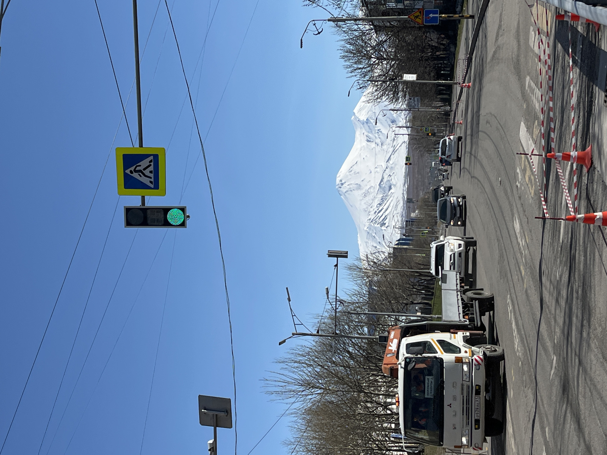 The image depicts a street scene in Petropavlovsk-Kamchatsky, Russia, characterized by a mix of natural and urban elements under a clear blue sky. In the background, a large snow-capped mountain dominates the horizon. In the foreground, there is a road with multiple vehicles, including a white flatbed truck with an orange boom, and other cars traveling on the right side of the road. Roadside infrastructure includes streetlights, bare trees lining the road, and traffic control devices. A green traffic light is active, and a crosswalk sign is visible. Safety cones and barricades delineate part of the road, potentially indicating construction or maintenance. The road surface appears to be asphalt, showing some wear and tear with visible tire tracks.