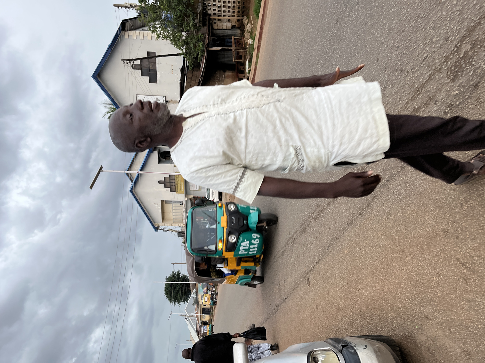 The image shows a man walking down a street. He is wearing a white, embroidered shirt and dark pants. His head is tilted back and his eyes are closed, with his mouth open. He appears to be mid-stride, with one arm extended forward. Behind him, a green and yellow auto-rickshaw is visible on the road. The auto-rickshaw has the license plate "PTA-11169" visible. In the background, there are buildings and trees. The sky is overcast, suggesting a cloudy day. The overall impression is of a person in a moment of perhaps ecstatic or relieved expression while moving along a street. There are other vehicles and people further down the road, indicating an urban or semi-urban environment. The ground is paved, and there are electrical wires overhead.