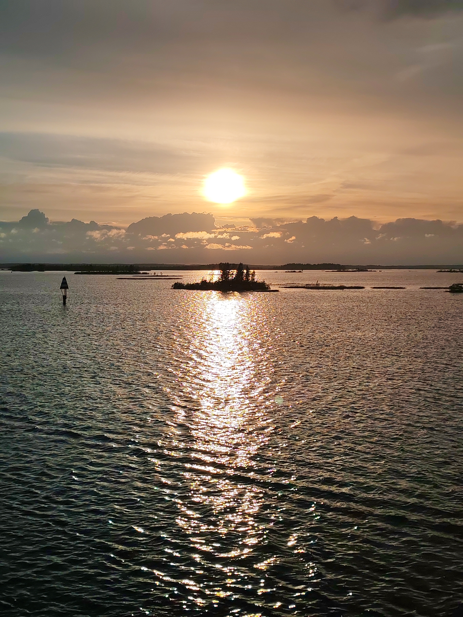 The image depicts a body of water, likely a lake or sea, at sunset. The sun is partially obscured by clouds, casting a golden glow across the sky and reflecting brightly on the water's surface. There is a small, tree-covered island in the middle ground and a navigational marker to the left. The water is rippled, and the overall scene is tranquil. There is no indication of the image location being Bereg, Russia. There are no people visible in the image.