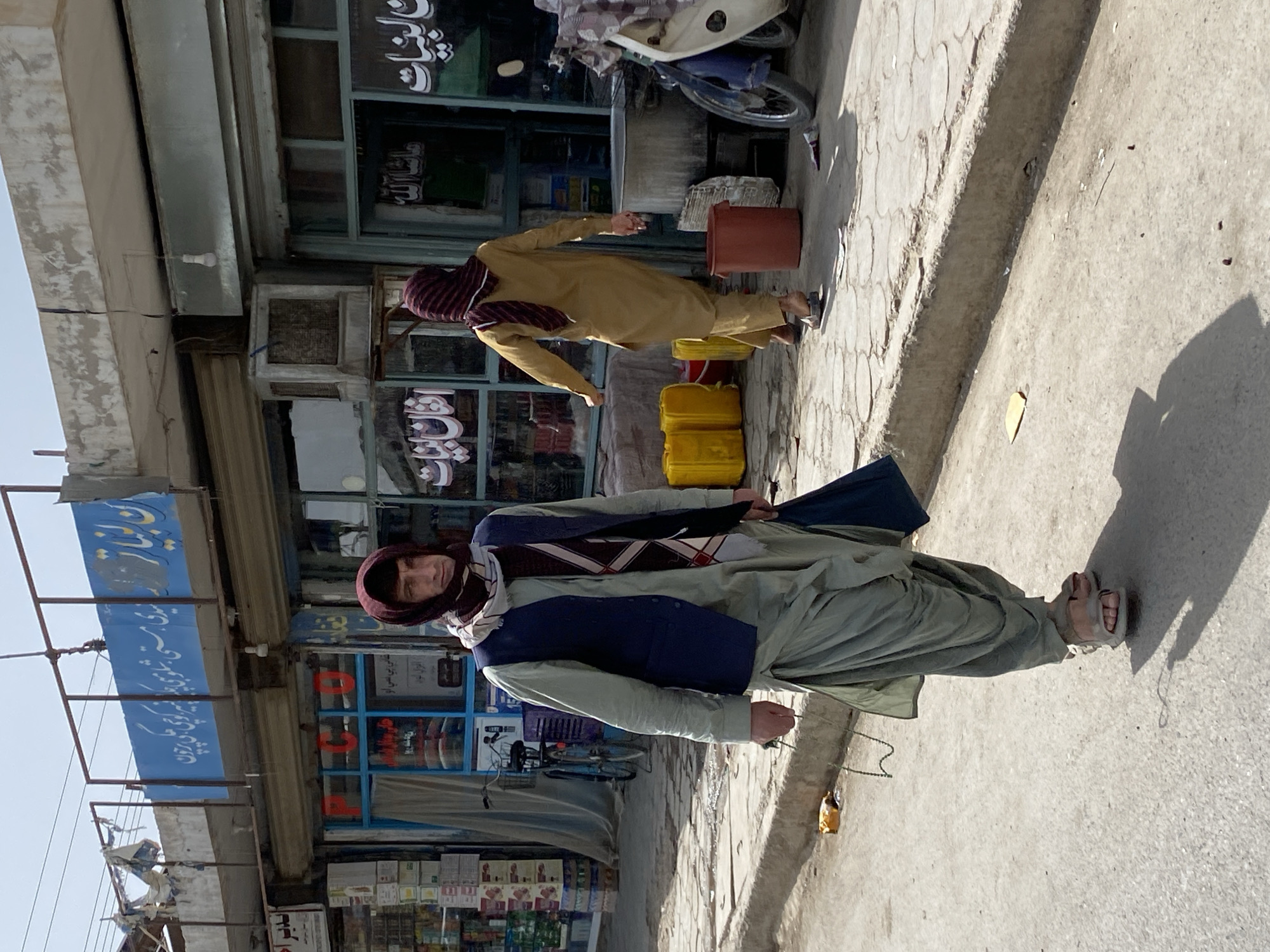 In Kandahar, Afghanistan, a man walks on a paved street in front of a row of storefronts. He is wearing a light green shalwar kameez, a dark blue vest, and a maroon and beige striped scarf wrapped around his head and neck. He carries a blue bag in his right hand and a rosary in his left. Another person in a tan shalwar kameez and maroon scarf is walking away from the camera to the right. The storefronts have glass windows and metal shutters, with signs displaying Arabic script. Yellow plastic containers are stacked near the entrance of one shop. A motorcycle is parked on the right side of the image. The street is worn and appears to be a commercial area.