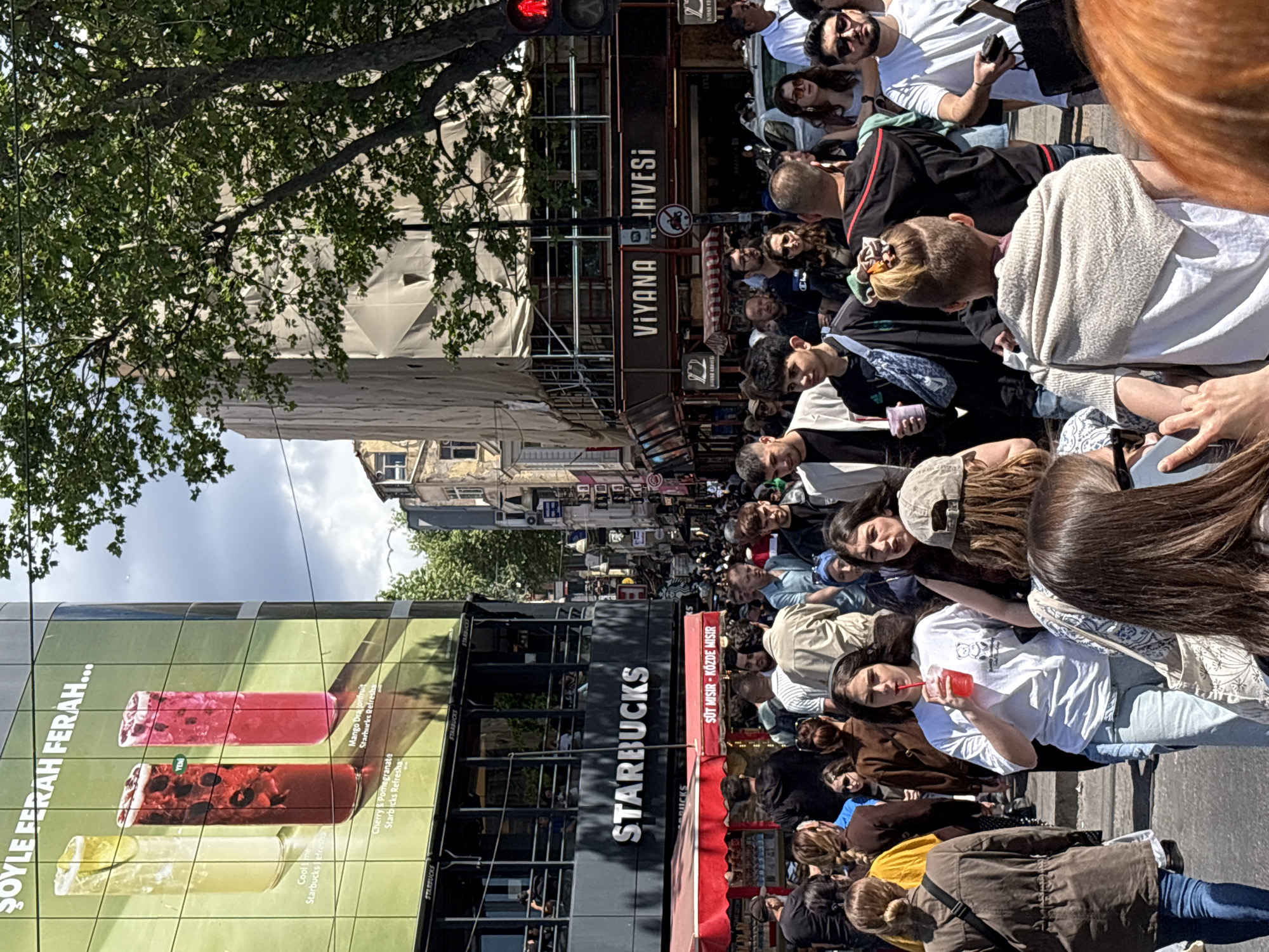 The image shows a crowded street scene in Kadıköy, Turkey. A large Starbucks store is visible on one side, featuring advertisements for Starbucks Refreshers. A second-story cafe, Viyana Kahvesi, is on the other side. Many people are walking or standing on the street, and some are drinking from cups. The overall impression is of a busy, urban environment.