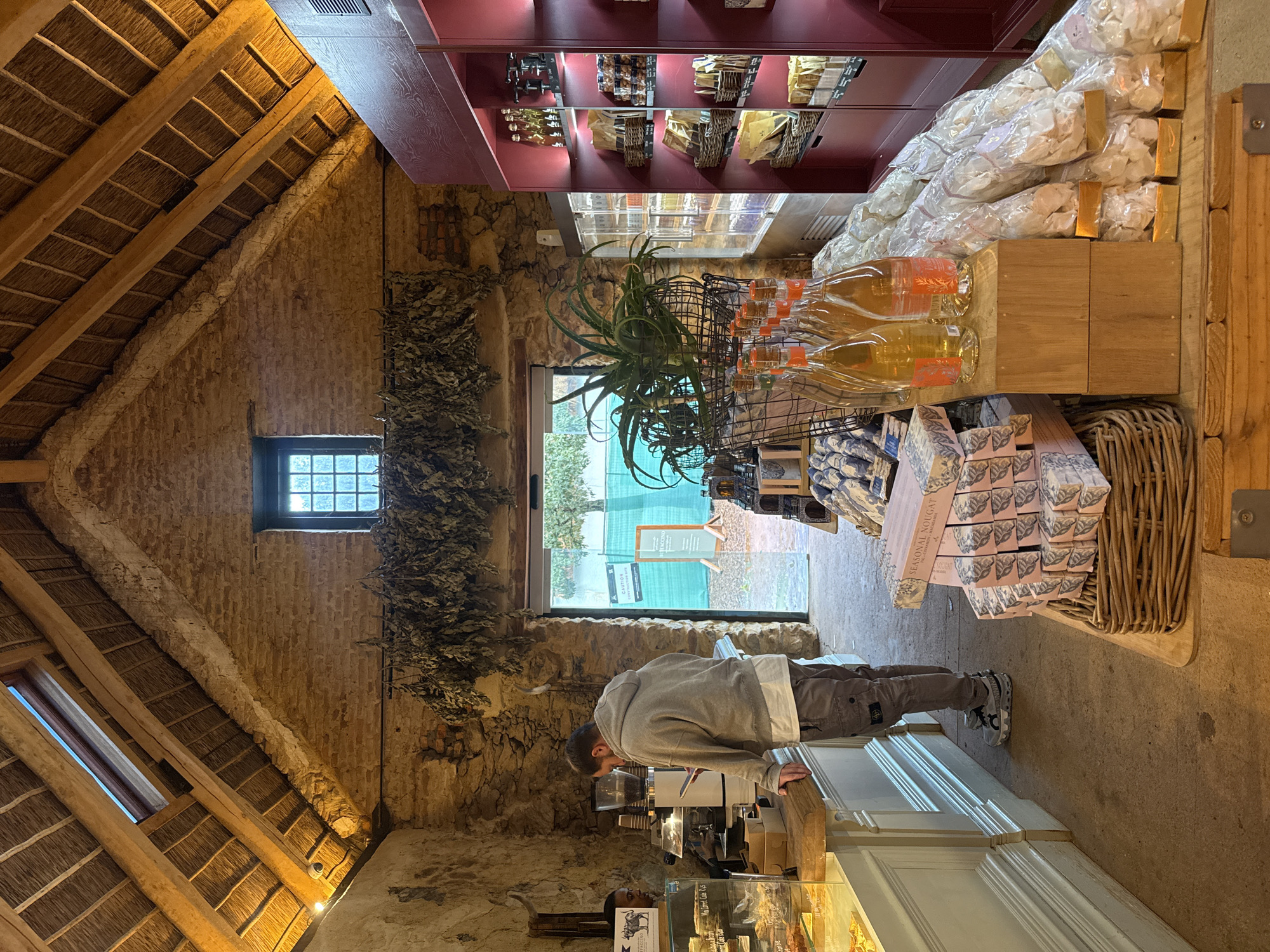 A man stands at a counter inside a rustic shop. The man is wearing a grey hooded sweatshirt, light brown pants, and grey athletic shoes. He is leaning forward towards the counter, where a coffee machine is visible.
The shop features exposed brick walls and a thatched roof held up by wooden beams. A window is set into the wall to the left of the man. Bunches of dried foliage hang near the window.
Shelves with products line the opposite wall. Items on display include bottled liquids, packaged nougat, and other goods. Through the doorway, a green screen and exterior elements suggest the shop opens to an outdoor area.
The shop is located in Drakenstein Nu, South Africa.