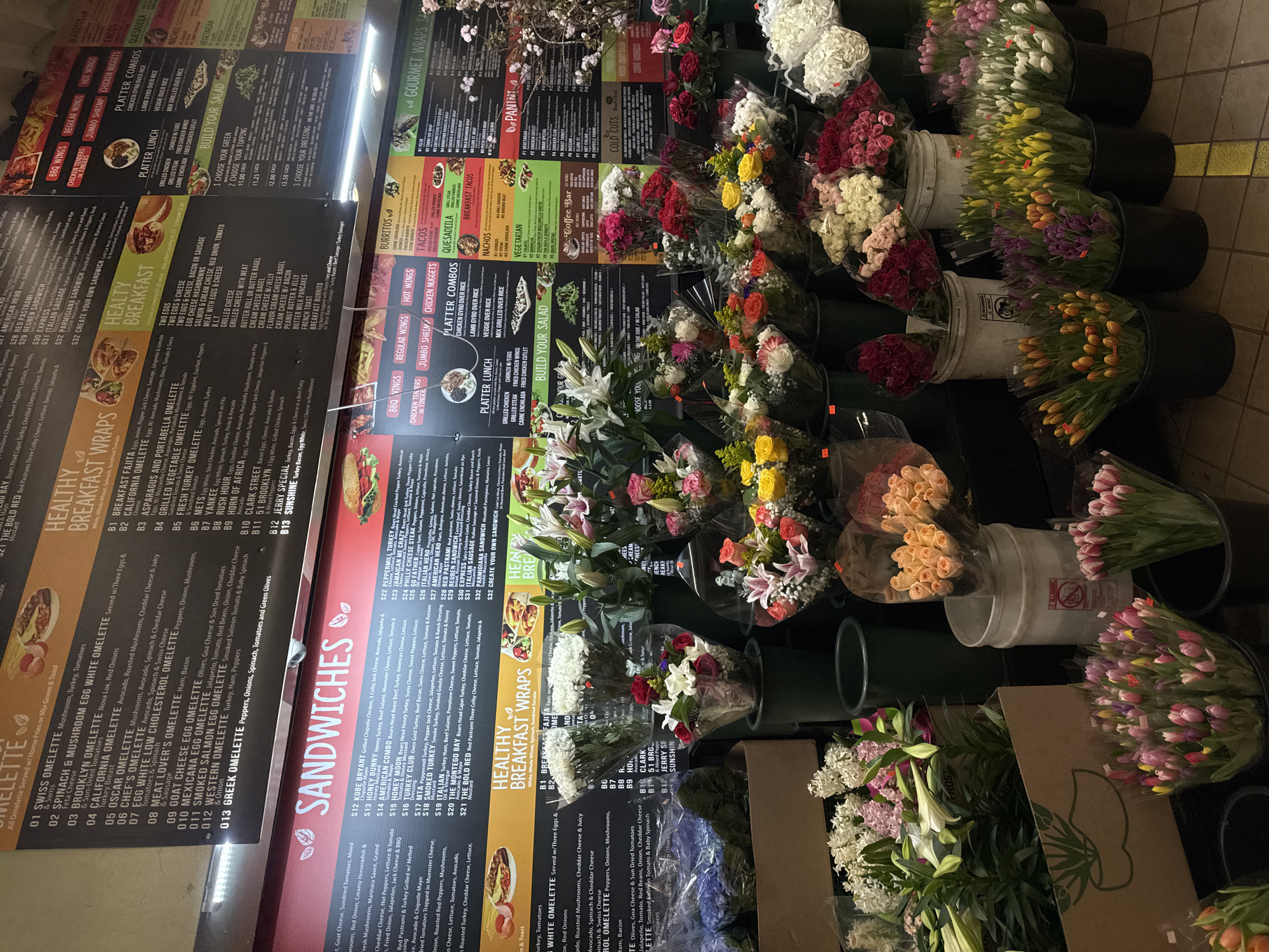 The image shows a deli or small grocery store in an urban environment with a large menu board advertising breakfast wraps, sandwiches, platters, and other food items. Bunches of fresh flowers are arranged along the wall. The location is likely New York City, based on menu item names like "Brooklyn Omelette" and "51 Brooklyn" and the overall urban aesthetic.