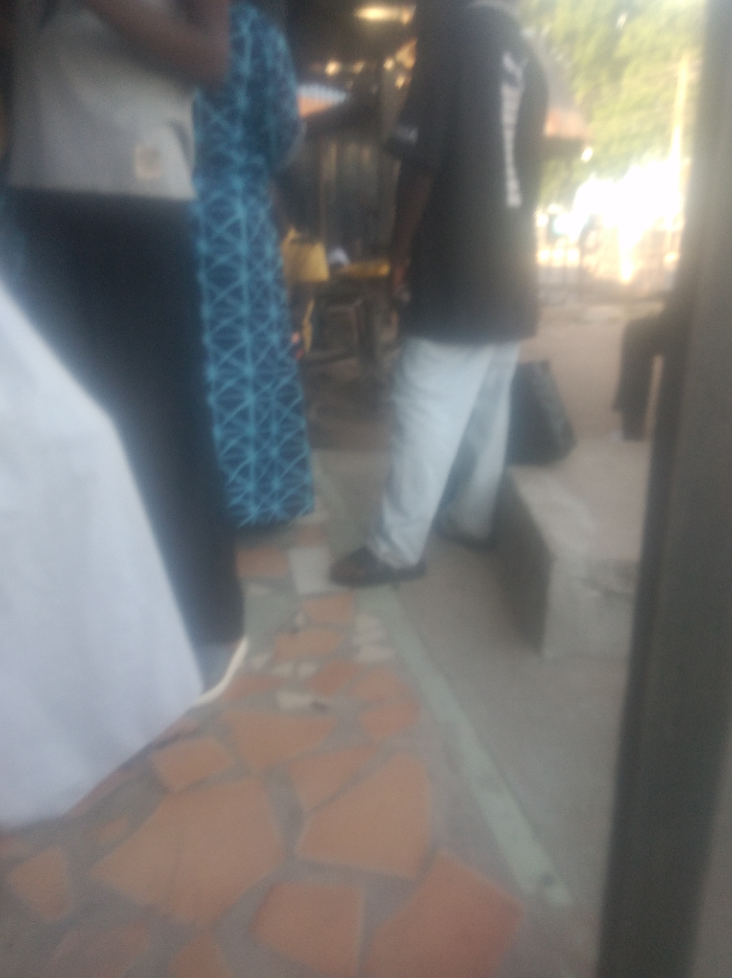The image depicts an outdoor scene with several individuals, likely in Jalingo, Nigeria. In the foreground, a person is visible from the waist down, wearing a white garment that obscures their lower body. To their right, another person is wearing a dark blue, patterned dress. Further to the right, a person in a black t-shirt with a white graphic on the back and light-colored pants is standing. Behind them, another individual, mostly obscured, is seen wearing dark clothing.
The setting appears to be an outdoor area with a tiled floor made of irregular, reddish-brown and grey tiles. In the background, there are structures that suggest a commercial or casual gathering place, possibly a street market or a roadside eatery. The lighting suggests it is daytime, with the sun casting a bright glow from the right side of the frame, creating some overexposure and blurring. The overall mood is indistinct due to the blurriness of the image. No discernible text is visible in the image. The activity appears to be people socializing or going about their day in a public space.