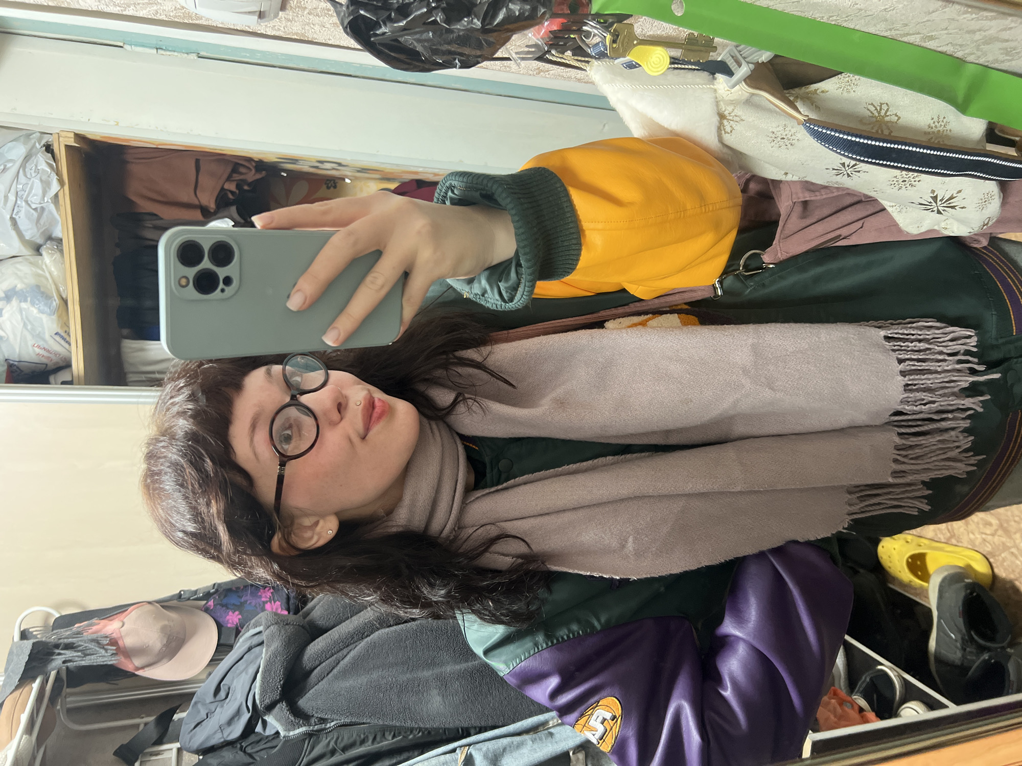 A young person takes a mirror selfie in a cluttered indoor setting. The subject is a female with dark brown, wavy hair and bangs, wearing round black-framed glasses. She has a small stud piercing above her lip and another on her earlobe, and a neutral, slightly posed expression while looking into the camera. She holds a light grey smartphone with a triple-lens camera array in her right hand, which is visible in the reflection.
She is dressed in a multi-colored jacket: primarily dark green, with vibrant yellow sleeves featuring ribbed dark green cuffs. A section of the jacket on her right side is purple, adorned with a yellow patch displaying the letters "LS" and a basketball logo. Around her neck is a light pinkish-grey fringed scarf, wrapped loosely.
The background, reflected in the mirror, reveals a personal and somewhat untidy room. Various items are visible: shelves containing bags and clothes, garments hanging on a rack (including one with gold snowflake patterns), keys, and a green strap. On the floor or a lower shelf, several pairs of shoes can be seen, notably a bright yellow pair and dark sneakers. Other clothing items, a brown hat, and a black jacket contribute to the crowded appearance of the space. The setting suggests a personal living space, likely a bedroom or dressing area, located in Chaikovsky, Russia. No specific time of day or weather is discernible from the indoor environment.