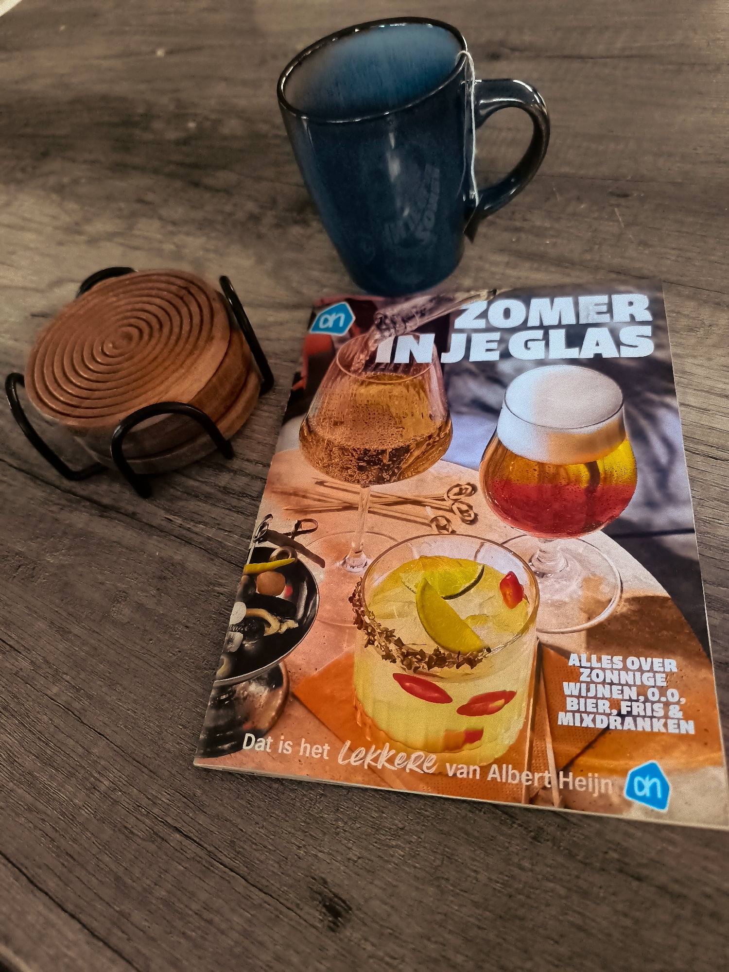 The image displays a top-down view of a wooden table with various objects. In the upper right quadrant, a dark blue ceramic mug is positioned, containing a white tag hanging from its handle. To the left of the mug, a stack of round, wooden coasters rests in a black metal holder. In the foreground, a magazine titled "ZOMER IN JE GLAS" is open, featuring images of drinks. The magazine is partially overlapped by the mug and coasters. The text on the magazine includes "ALLES OVER ZONNIGE WIJNEN, O.O. BIER, FRIS & MIXDRANKEN" and "Dat is het Lekkerste van Albert Heijn". The overall lighting suggests an indoor setting. There are no people or animals visible in the image, and no specific location in Almere, Netherlands can be determined.