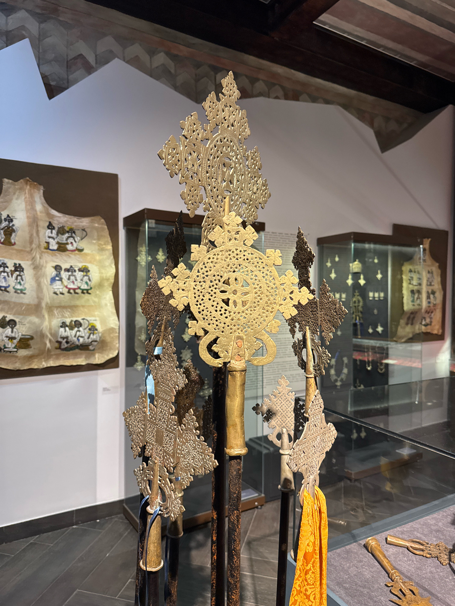 The image shows a display of Ethiopian crosses inside a museum. The crosses are made of metal, some gold-colored and others darker, and are intricately designed with geometric patterns. They are mounted on poles, with one cross having an orange fabric streamer attached. In the background, glass display cases contain more crosses and other religious artifacts. To the left of the central display is a framed artwork depicting figures in traditional clothing. The museum walls are white, and the floor is dark grey. The ceiling has exposed wooden beams and decorative patterns. There are no people visible in the image. The exact location in Moscow, Russia is not provided in the image.