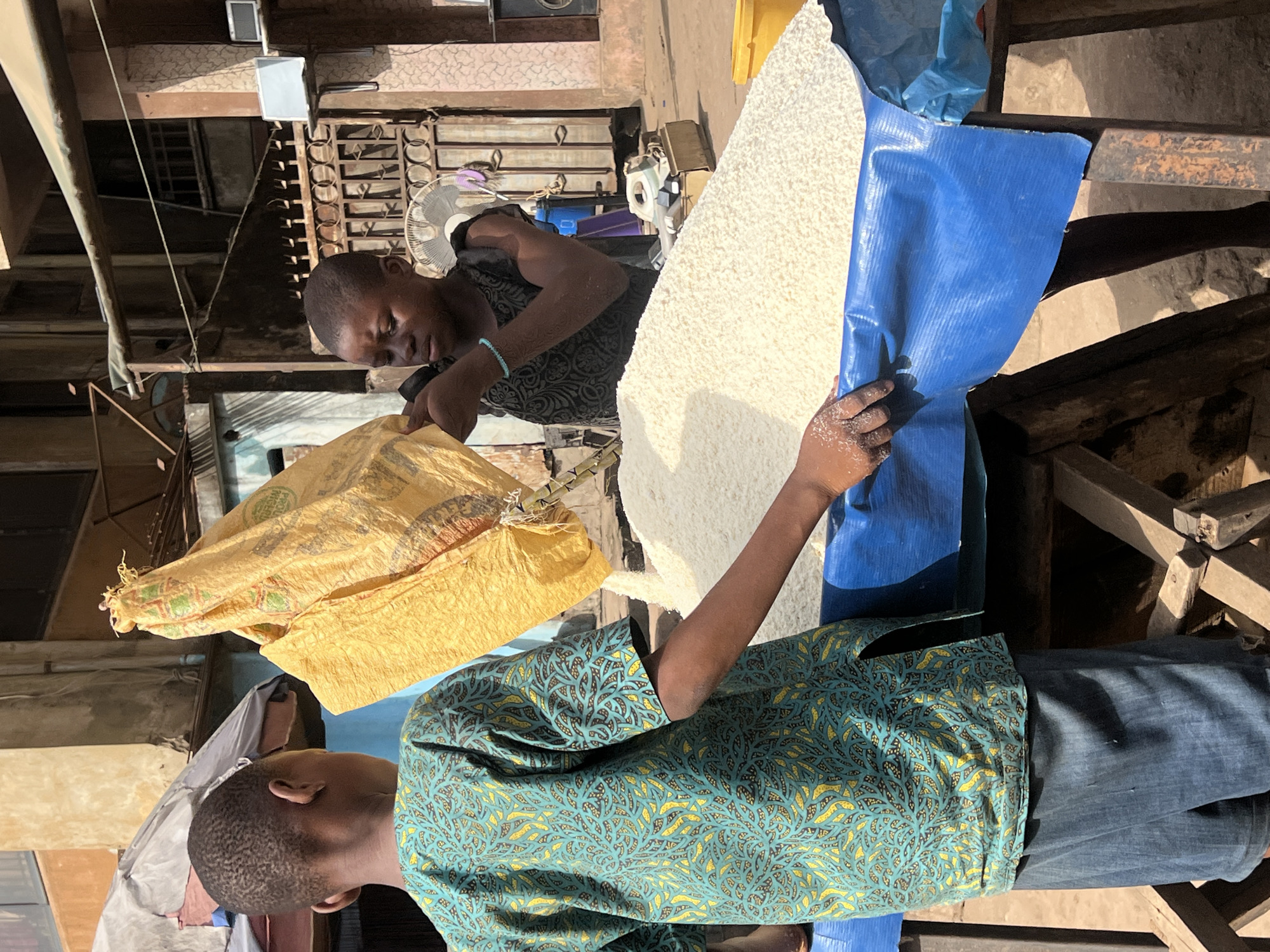 This image, captured in Nkpor, Nigeria, depicts an outdoor commercial scene involving two young individuals handling a large quantity of a white granular substance, likely cassava flakes (gari), a common staple food.
In the foreground, a person, seen from the back, wears a distinctive blue-green shirt adorned with yellow and brown leaf-like patterns and blue jeans. This individual uses their right hand to hold and manage a blue tarp which contains a significant mound of the white granular material. Traces of this substance are visible on their hand.
Positioned behind and slightly to the right, another individual, wearing a dark patterned shirt and a light blue bracelet on their left wrist, is actively engaged in the process. They are holding open a large, crumpled yellow sack, seemingly in preparation to fill it or having just filled it with the white granular material. Their hands are also covered with the substance, indicating direct handling.
The white granular material is spread out on the blue tarp, suggesting it is being portioned, weighed, or prepared for sale. The background reveals an urban or semi-urban setting with multi-story buildings, featuring visible balconies, metal railings, and a wall-mounted fan. The bright, natural lighting indicates it is daytime, with clear shadows cast. The scene conveys a sense of daily market activity. No discernible text is clearly visible on the sacks or in the surroundings.