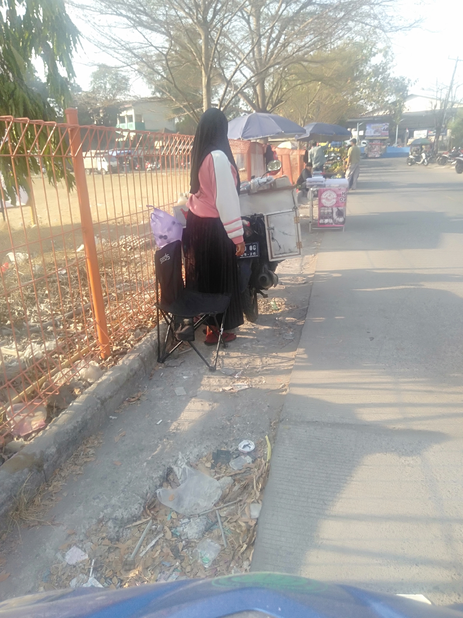 A woman wearing a hijab and a pink and white jacket is standing next to a motorcycle and a folding chair on the side of a road in Sukadami, Indonesia. The motorcycle has a license plate that reads "B 8548 OG". A purple bag is placed on the seat of the folding chair. In the background, there are several food carts and umbrellas. The scene appears to be a street market. The ground is littered with trash and dry leaves. The sky is clear and the sun is shining.