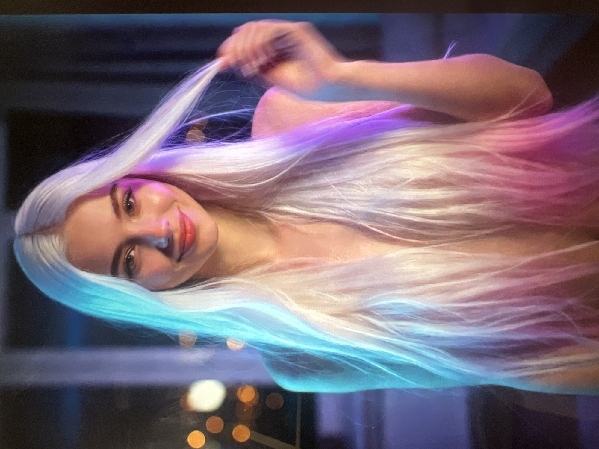 A woman with long, platinum blonde hair is looking at the camera and smiling. Her hair has pink and blue highlights. She is wearing a light-colored garment that exposes her chest and shoulders. The background is dark with blurred lights. The image is lit with blue and pink lights. The scene appears to be indoors.