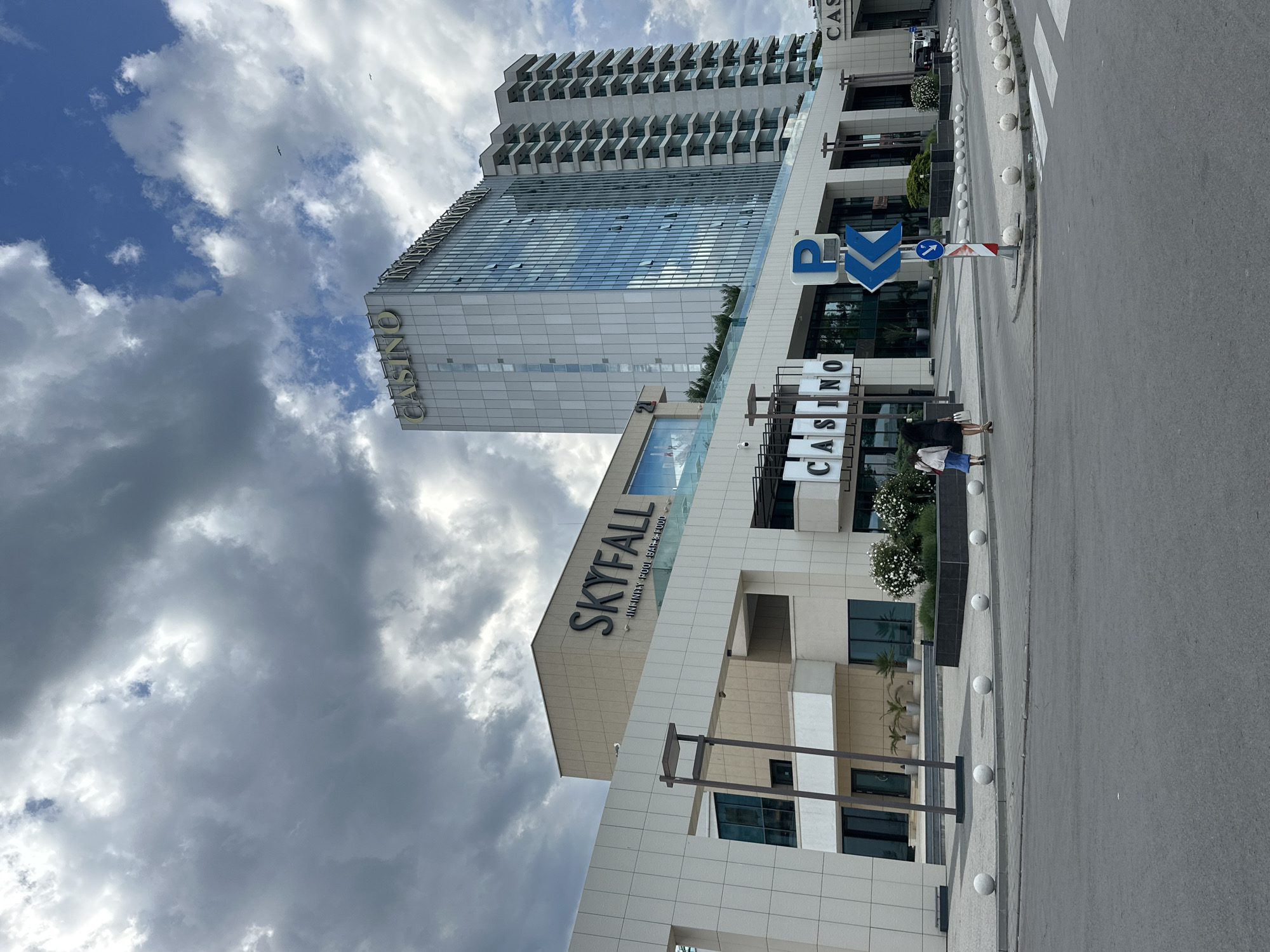 The image shows the Skyfall Casino and International Hotel Casino in Varna, Bulgaria. The Skyfall Casino is a modern, low-rise building with the name "SKYFALL" and "INFINITE POOL BAR & FOOD" visible on the facade. Next to it, the International Hotel Casino is a taller building with multiple balconies and "CASINO" written vertically on the side. A parking sign with a blue "P" is visible. On the street are two women and a row of spherical bollards. The sky is cloudy with blue patches.