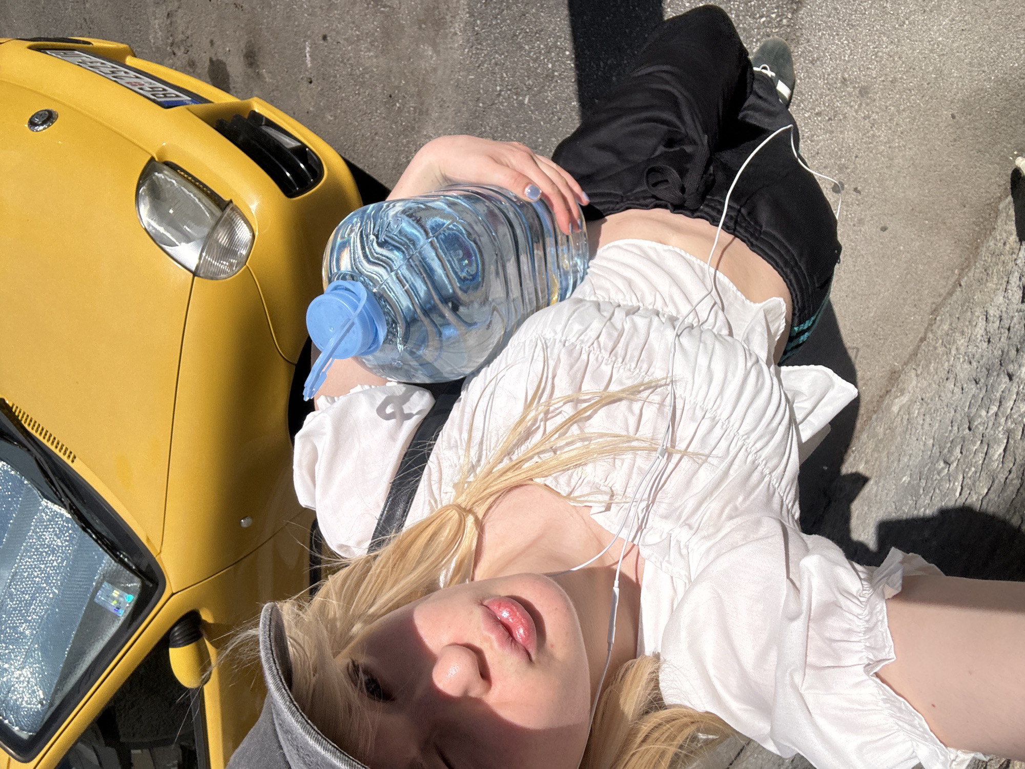 The image shows a person lying on their back outdoors, with a yellow vehicle and a cobblestone surface visible. The person has blonde hair and is wearing a white ruffled top and black pants. They are holding a large bottle of water, and wearing white earbuds. Their face is tilted upwards, and their lips are parted. The sun is shining brightly, casting shadows and creating a high contrast lighting. The scene appears to be in an urban setting, possibly a street or sidewalk, given the presence of the vehicle and pavement. The time of day is likely midday or early afternoon due to the strong sunlight and shadows. The person's relaxed pose and the act of holding water suggests they might be resting or hydrating after some activity. The overall mood is casual and relaxed, with a focus on the subject and the immediate surroundings. There is text visible on the license plate of the yellow vehicle, but it is not legible.