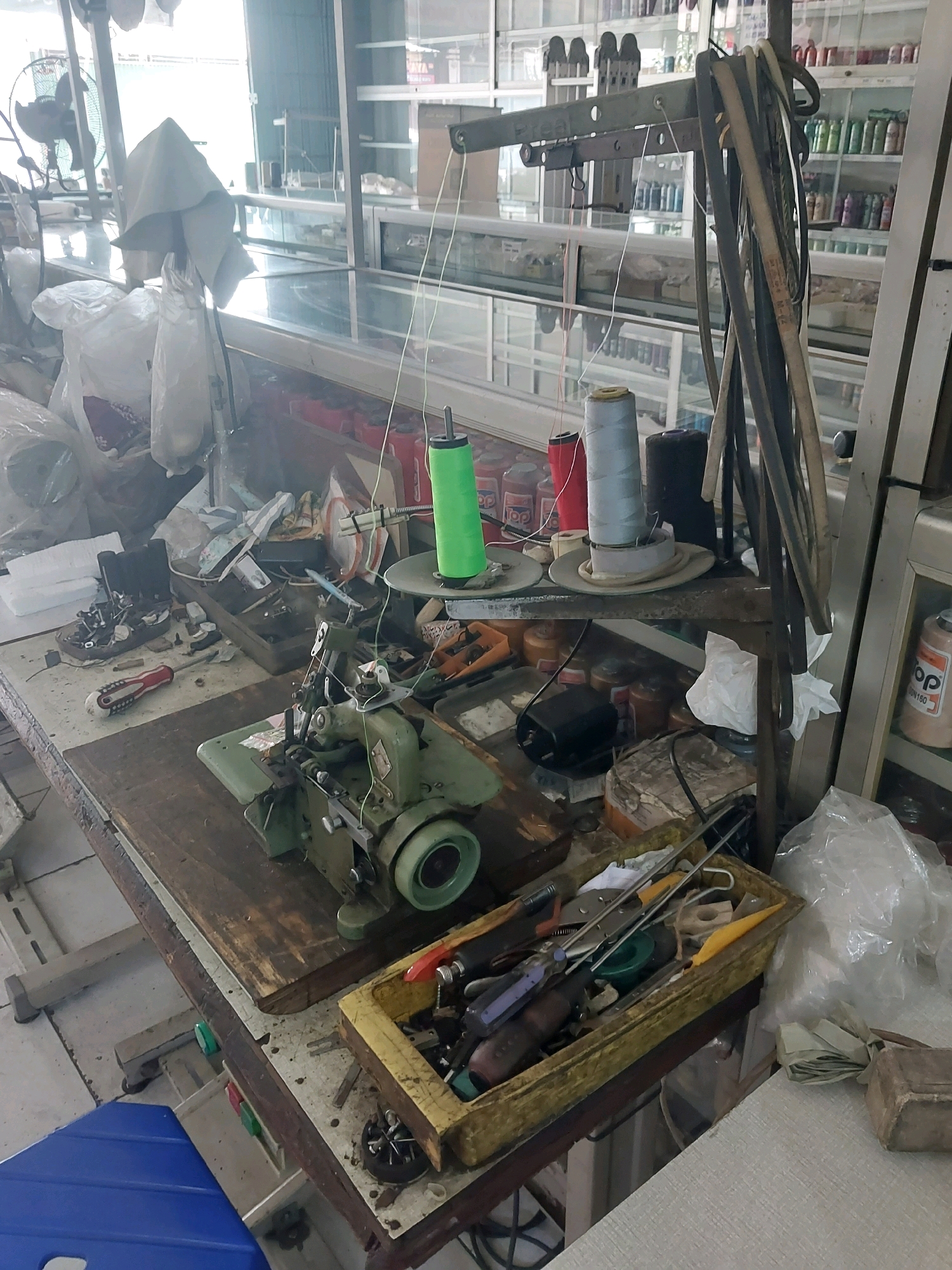 The image depicts a workshop, likely a sewing or tailoring establishment, situated in Malang, Indonesia. The foreground is dominated by a workbench laden with sewing equipment and various tools. A green industrial sewing machine is prominent, with threads of different colors, including green and light blue, running from spools above. Various tools, such as pliers, screwdrivers, and scissors, are organized in a yellow tray on the workbench. Rolls of fabric or other materials are visible in the background, some wrapped in plastic. Shelving units filled with spools of thread and other items are also present. A ceiling fan is visible in the upper left corner of the image. The overall environment appears functional and somewhat cluttered, indicative of an active workspace.