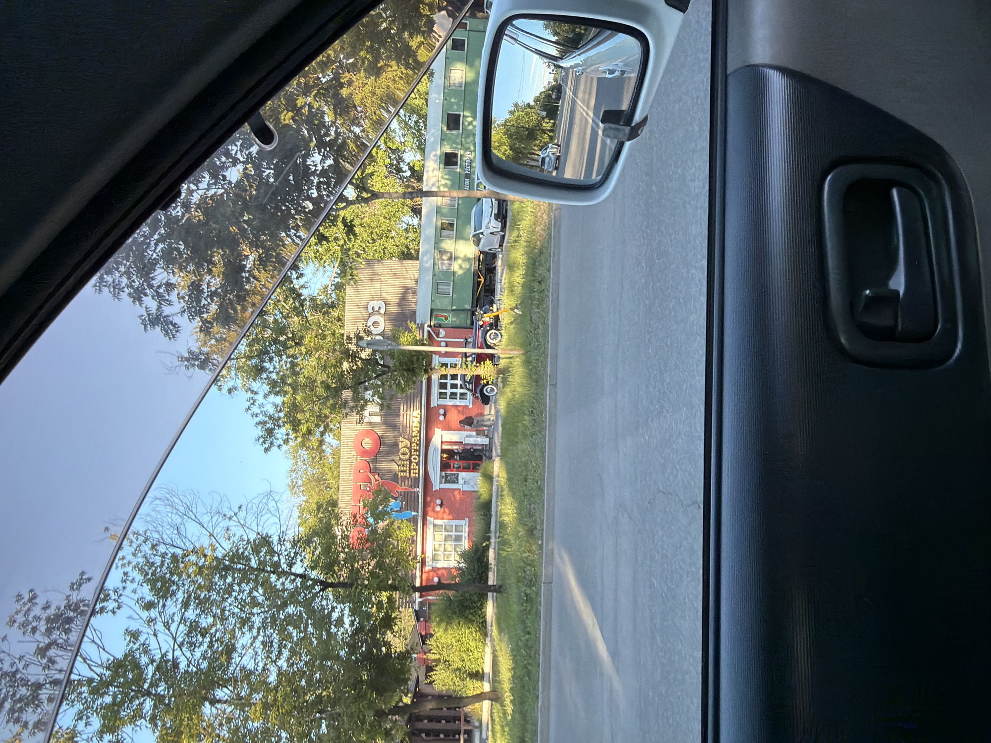The image, taken from the perspective of a car passenger, shows a street scene in Khabarovsk, Russia, on a sunny day. In the foreground, the interior of a car's door panel is visible. Through the car window, a red building with the word "ШОУ" (Show) in large letters is seen. Below this, smaller text reads "ПРОГРАММА" (Program). Several people are standing near the entrance of the building. To the right of the building is a long, green train car, identified by the text "ВАГОН РЕСТОРАН" (Restaurant Car) written on its side. The train car appears to be stationary, adjacent to the building and a grassy area. Trees line the street, and the sky is clear and blue. The reflection in the car's side mirror shows a street with other vehicles and buildings. The overall impression is an outdoor commercial establishment with a unique theme, possibly related to entertainment or dining, located in an urban environment.