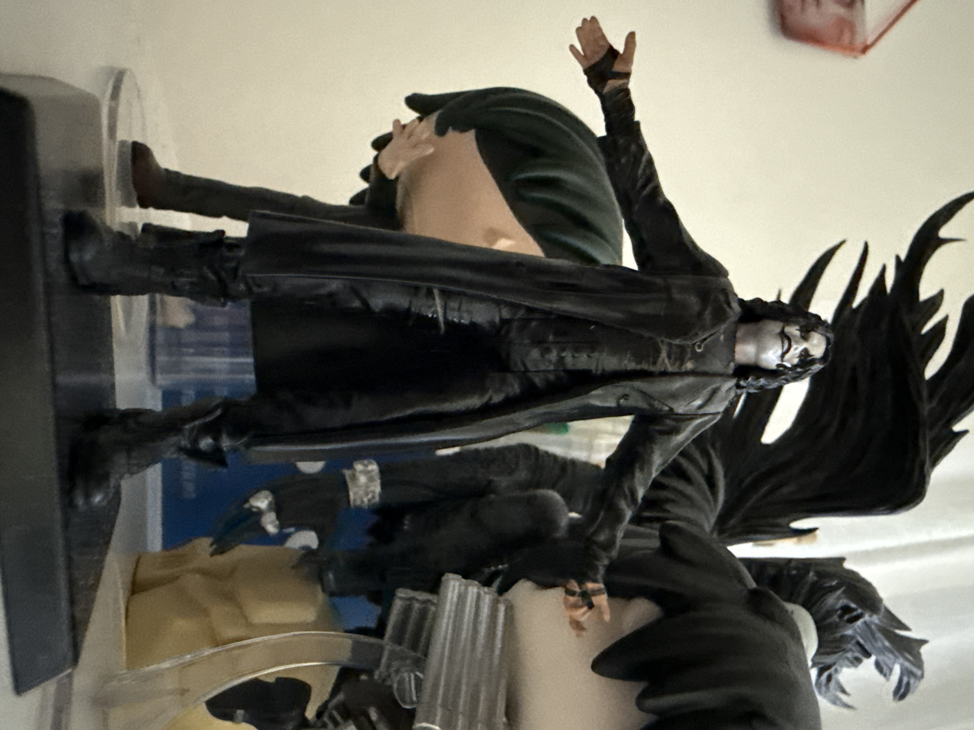 A close-up, eye-level, full shot captures a figurine of the character "The Crow" standing with arms outstretched. The figurine is dressed in a long black trench coat over a black suit, black boots, and black gloves. The character has dark, wavy hair and white face paint with dark eye makeup and dark lipstick. Behind "The Crow" are two other figures: one with dark green hair and a light-skinned face with a hand covering its mouth, and another figure with large black wings and black hair. The scene is set indoors against a light-colored, plain background. The figurine is positioned on a dark platform, with other objects, including what appears to be a metallic prop gun and a light brown, textured object, visible in the background. The scene does not depict any elements related to the city of Bad Sachsa, Germany.