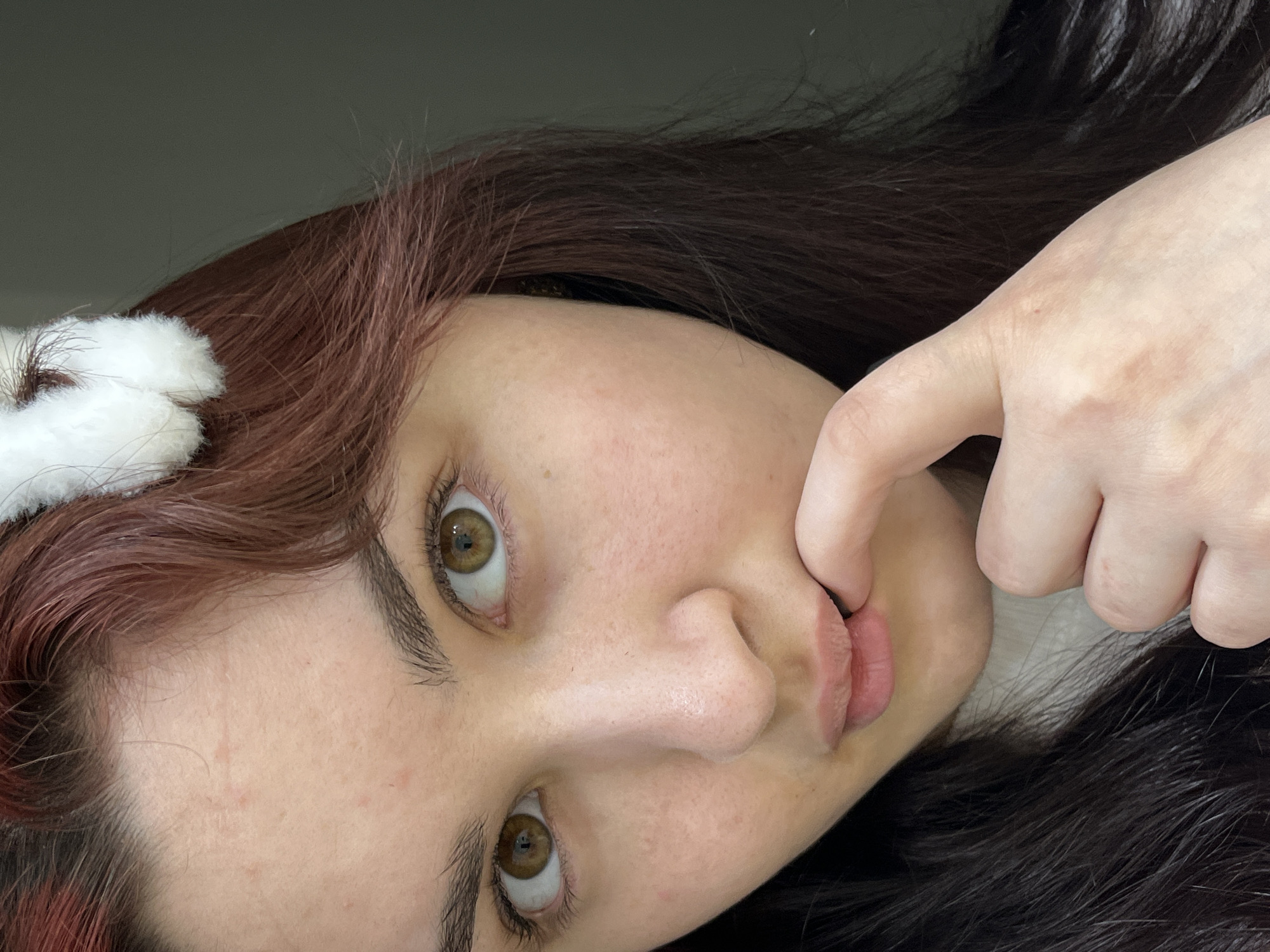 A close-up photograph shows a person's face. The person has fair skin, brown hair with red highlights, and brown eyes. They are wearing a white, furry accessory in their hair. One of their fingers is touching their lips, causing them to pout slightly. The background is a plain, light gray. The image does not contain information regarding the city of Neftekamsk, Russia.