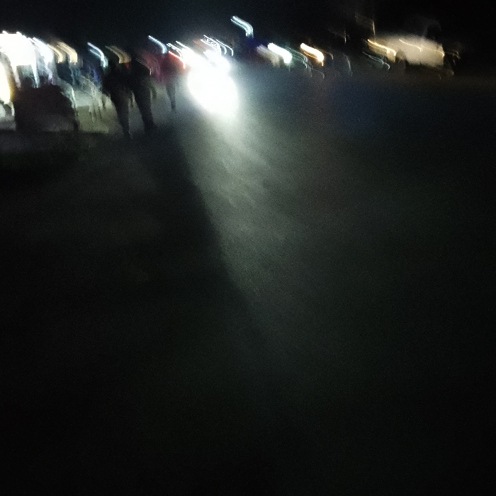 A nighttime street scene displays blurred figures walking on the left. A vehicle's headlights illuminate a path in the center. Light sources flicker in the background, possibly from streetlights or buildings. The overall impression is of motion and limited visibility due to darkness.